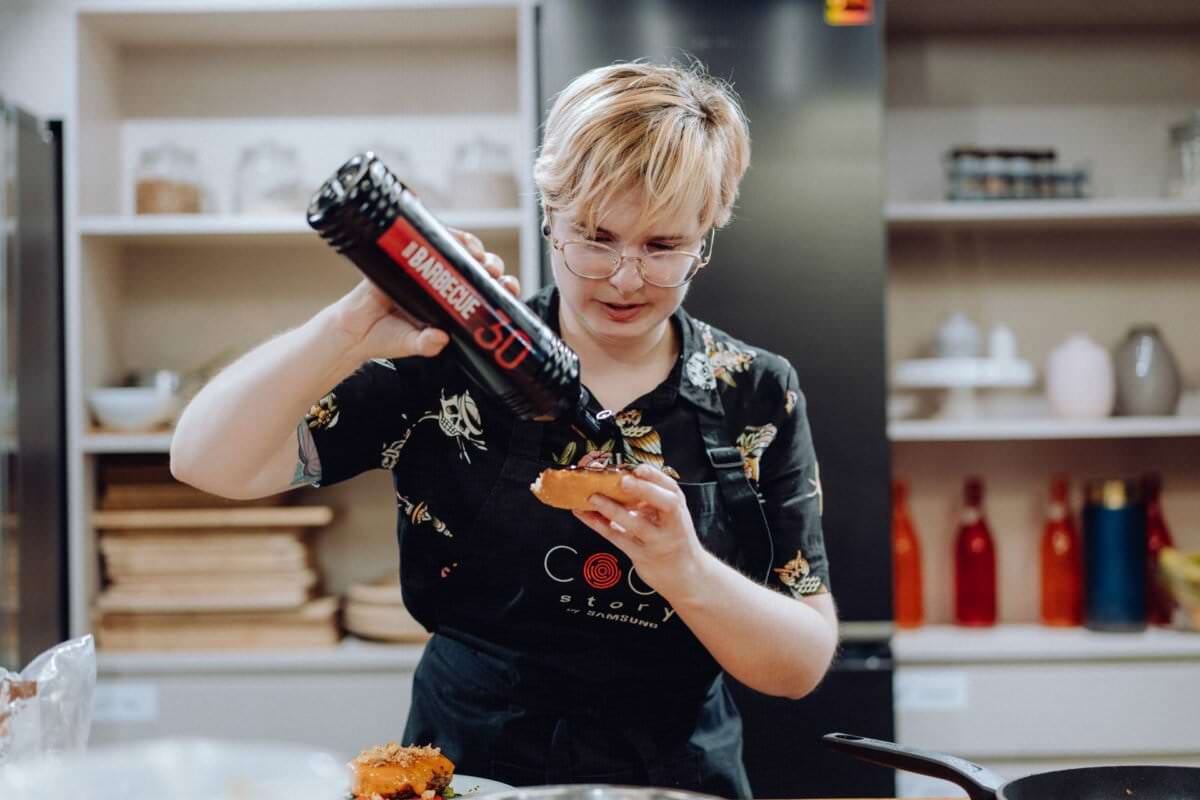 Osoba o krótkich blond włosach w okularach i czarnym fartuchu ostrożnie wyciskającą butelkę sosu barbecue na jedzenie w nowoczesnej kuchni. W tle widać półki z różnymi przedmiotami, które uchwycą autentyczny moment, idealny na sesję zdjęciową eventową.