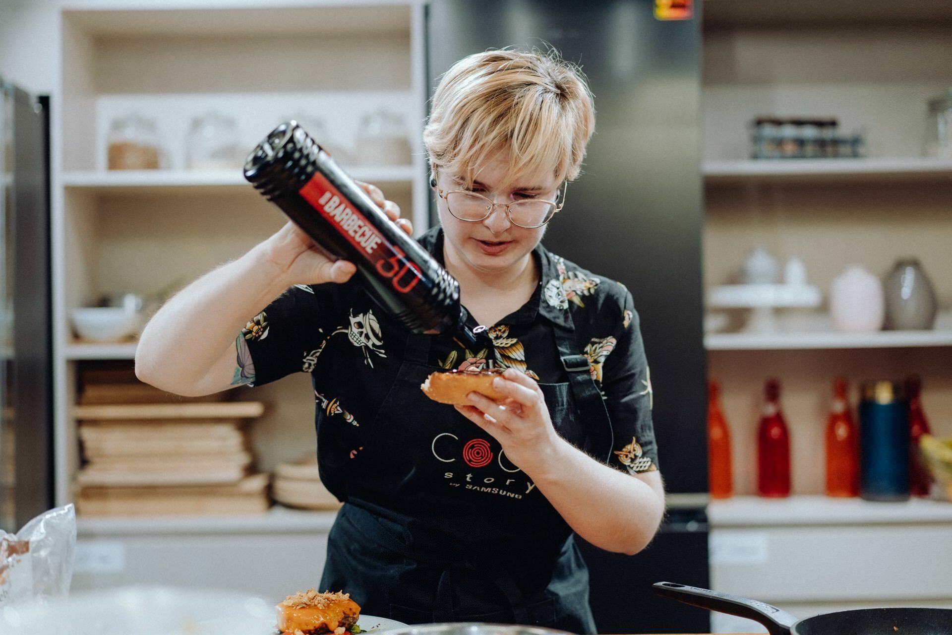 A person with short blond hair wearing glasses and a black apron carefully squeezing a bottle of barbecue sauce onto food in a modern kitchen. In the background you can see shelves of various items that capture an authentic moment, perfect for an event photo shoot. 