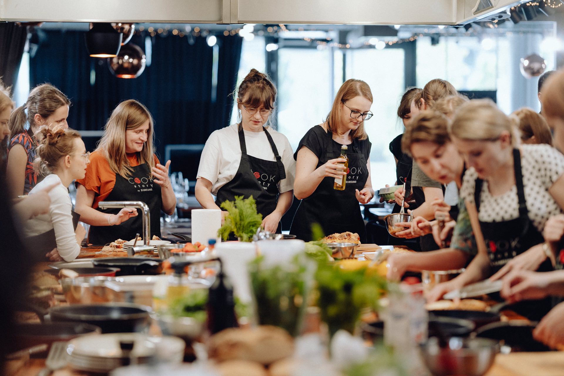 A group of people in aprons are busy preparing food around a large kitchen island. Some are chopping vegetables while others are mixing ingredients or discussing recipes. The atmosphere seems lively and collaborative, with various kitchen utensils and fresh ingredients scattered around - a perfect scene for an event photographer.  