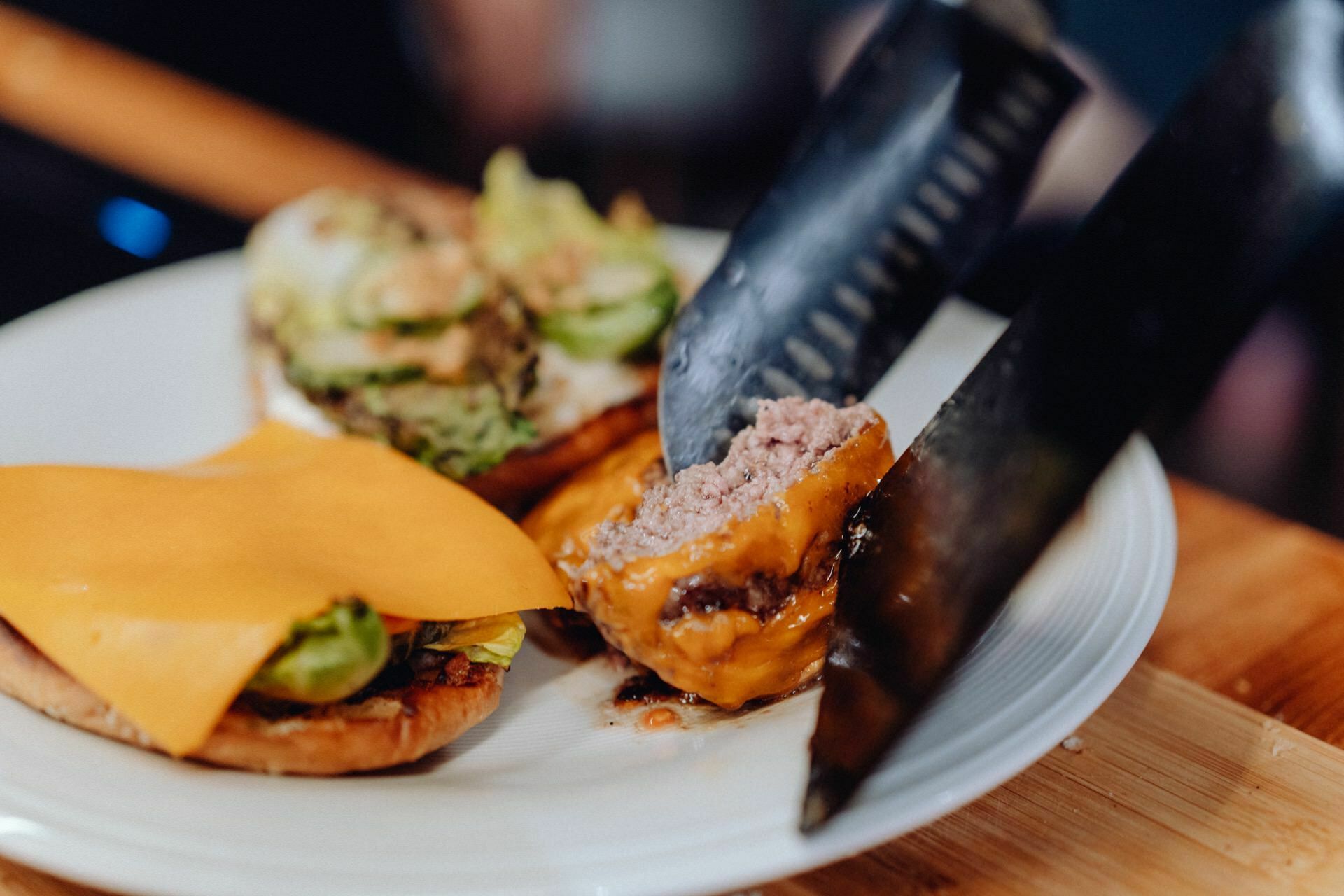 Close-up of a partially assembled cheeseburger on a white plate. A knife cuts through the burger, revealing its pink interior. The sandwich consists of a patty with melted cheese, lettuce and other ingredients in a bun. Perfect for any event photo shoot or event photography.   