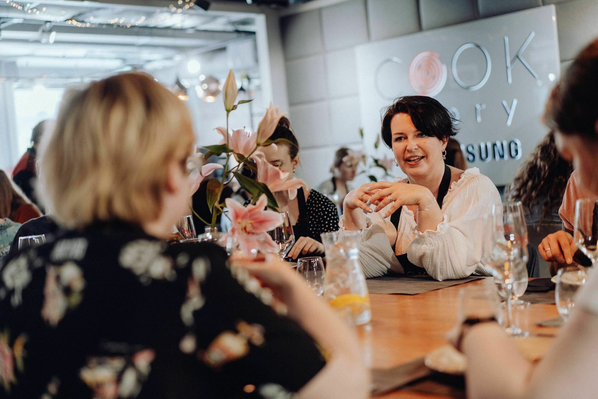 A group of people sitting around a wooden table having a conversation during a social event. In the center of the painting is a woman in a white blouse, smiling and gesturing with her hands. Flowers and glasses are lying on the table, and in the background is a sign that reads "READ". This could be a perfect example of event photography involving capturing vivid moments.   