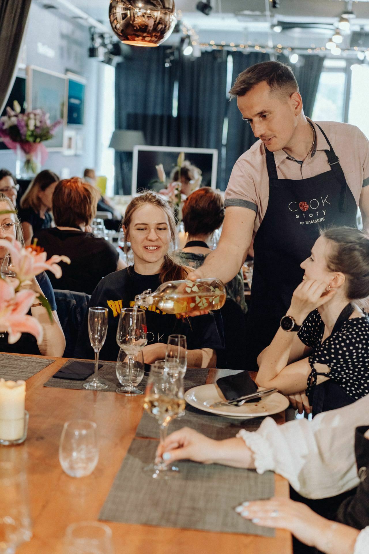 A man in an apron pours wine for a smiling woman seated at a table during a cooking class. The other participants, also seated, are watching and talking. The room is decorated with flowers, and various kitchen utensils can be seen in the background - a perfect scene for any Warsaw event photographer to capture event photo coverage.  