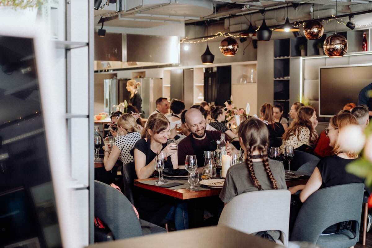 Tętniąca życiem scena restauracyjna z ludźmi jedzącymi i rozmawiającymi przy kilku stołach. Wystrój obejmuje nowoczesne elementy z metalowymi akcentami i przyćmionym oświetleniem. Na stołach ustawiono talerze, szklanki i sztućce. Na pierwszym planie dwie osoby zajadają się posiłkiem i piciem, co zostało uchwycone idealnie na potrzeby fotorelacji z wydarzeń.