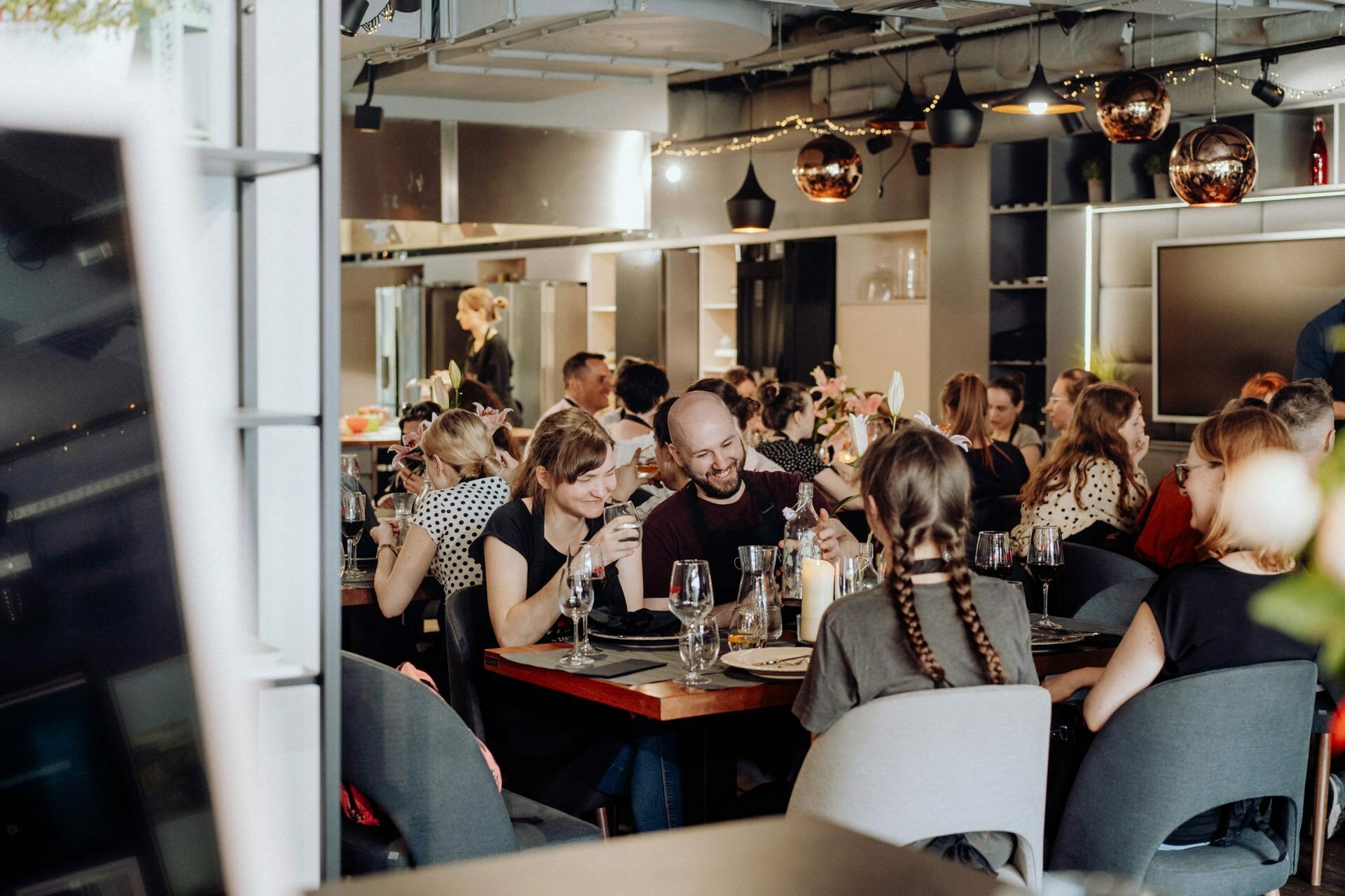 A lively restaurant scene with people eating and chatting at several tables. The decor includes modern elements with metal accents and dim lighting. Plates, glasses and cutlery are set on the tables. In the foreground, two people are enjoying a meal and a drink, which was captured perfectly for the photo coverage of the events.   