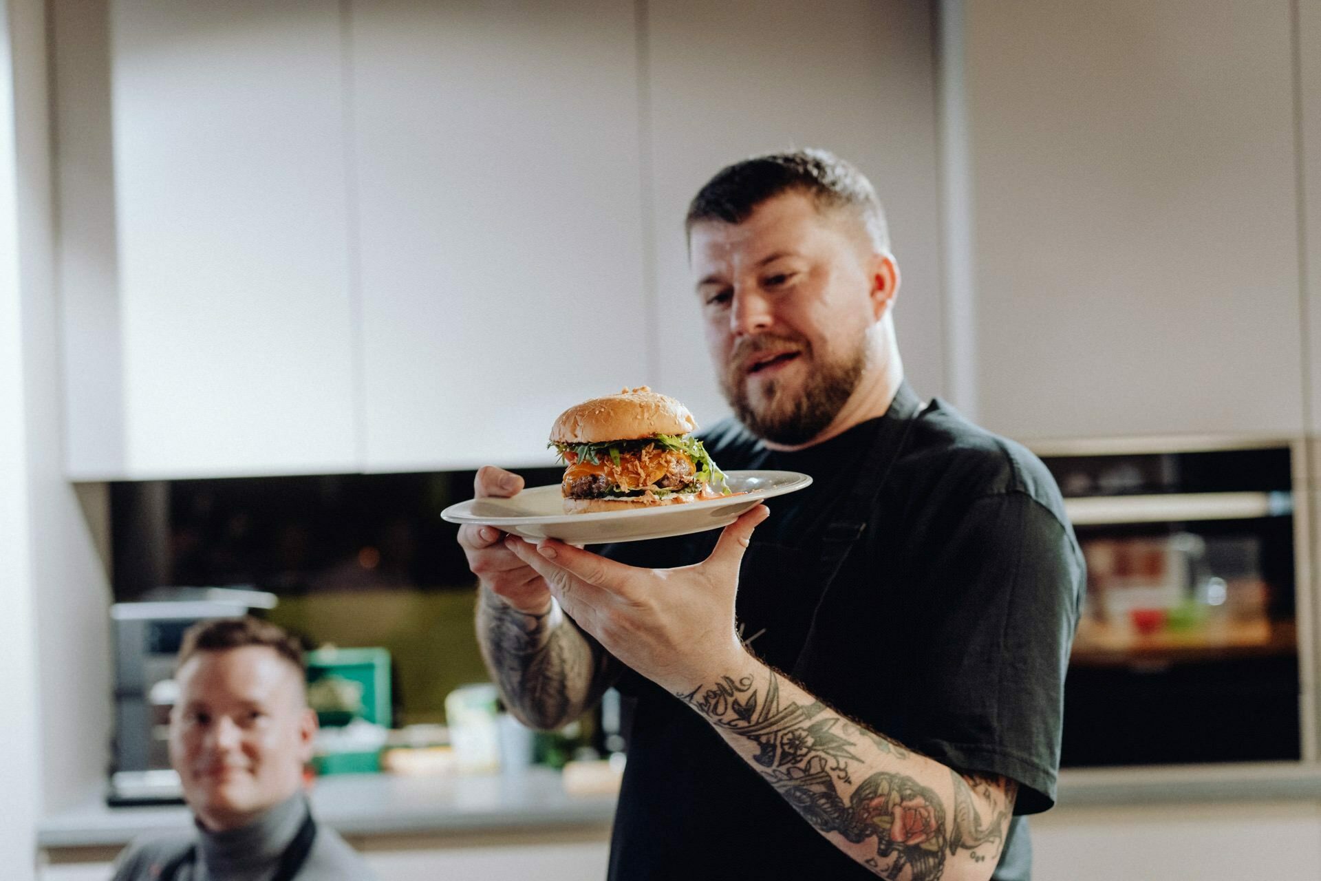 A man with tattoos on his arms proudly holds a plate with an exquisite burger in a modern kitchen with bright cabinets. In the background, another person sits and smiles, capturing the joy of the moment in this event photo. 