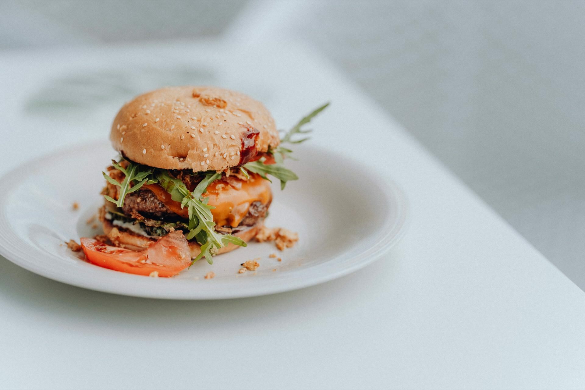 An exquisite burger with a sesame seed bun, arugula, cheese, a slice of tomato and a meat cutlet sits on a clean white plate. Crumbs and a piece of tomato are scattered all over the plate. The background is blurred, highlighting the burger - perfect for your event photography needs.  