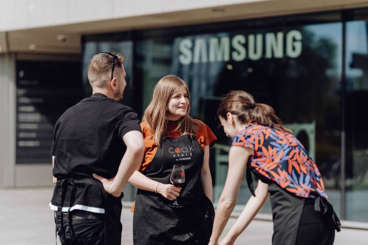 Trzy osoby w fartuchach prowadzą ożywioną rozmowę przed budynkiem z dużym szyldem Samsunga. Jedna osoba trzyma kieliszek wina, a dwie inne dzielą się beztroską chwilą, lekko pochylając się i śmiejąc. Otoczenie wydaje się swobodne i przyjazne, doskonale uchwycone na tej fotografii eventowej.