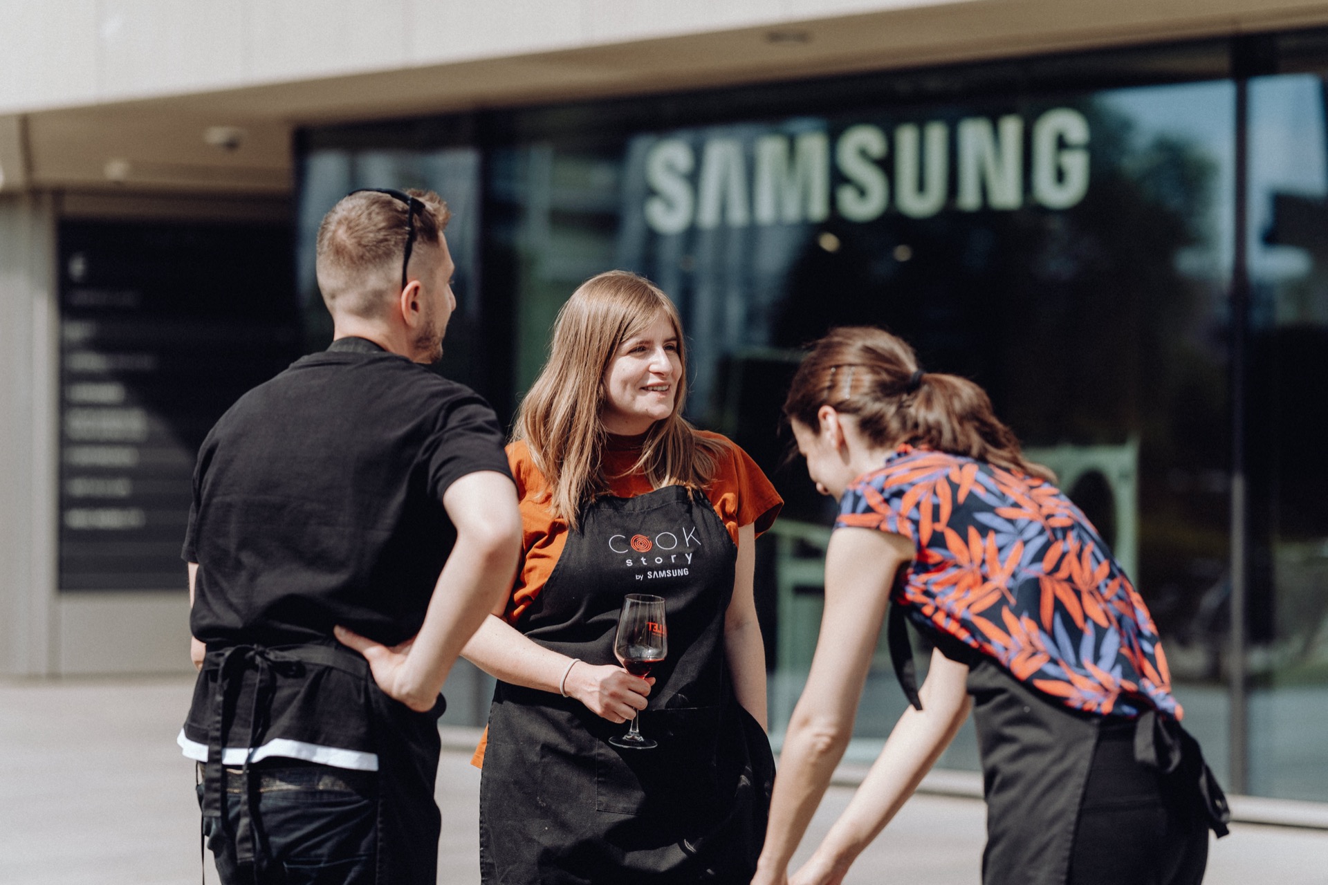 Three people in aprons are having a lively conversation in front of a building with a large Samsung sign. One person is holding a glass of wine, and two others are sharing a carefree moment, leaning slightly and laughing. The environment seems casual and friendly, perfectly captured in this event photography.  