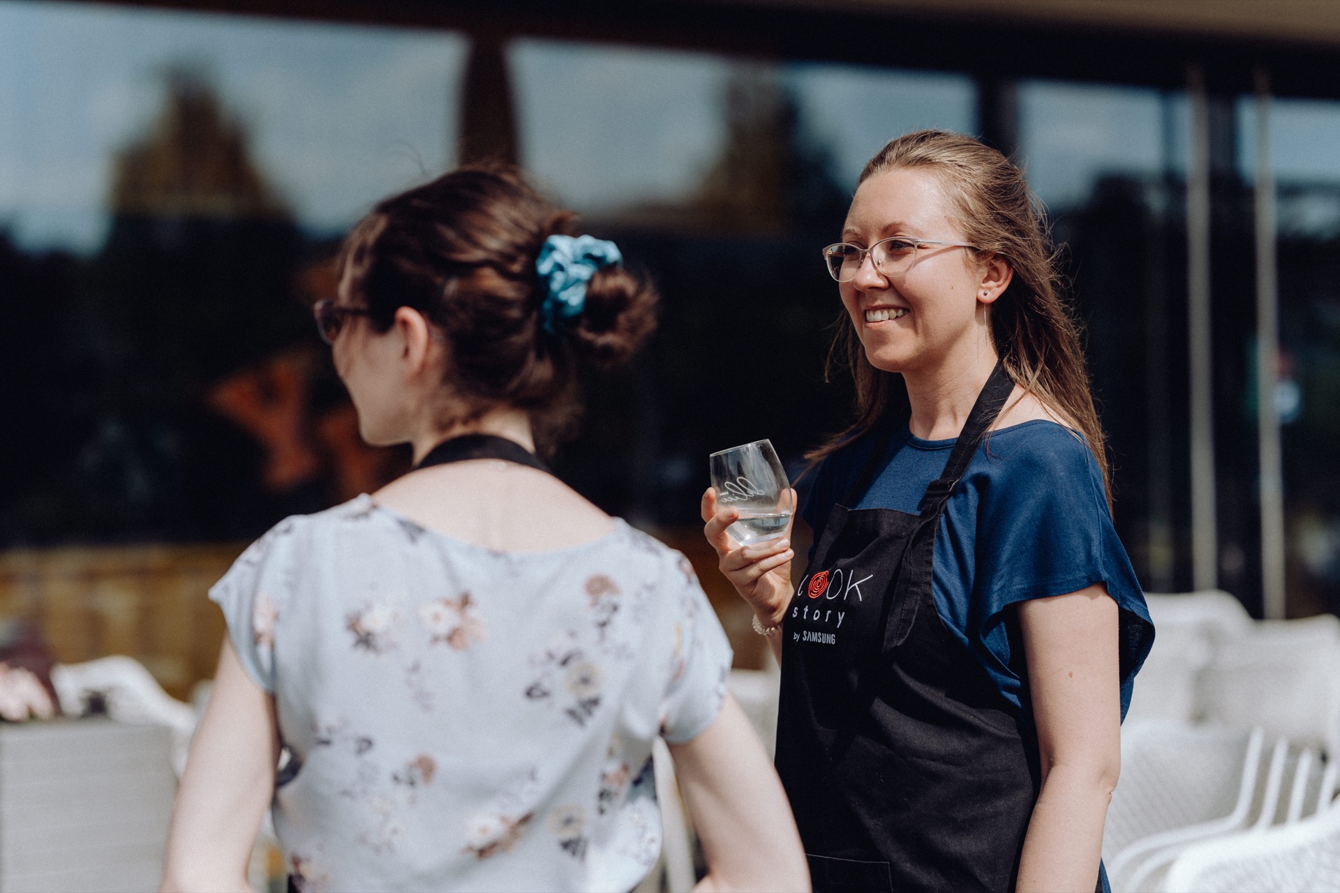 Two women are talking outdoors, one in a blue floral dress and the other in a blue top and apron with the word "COOK" written on it. The woman in the apron is holding a glass and smiling. They are standing in front of a building with large windows, surrounded by white chairs, beautifully captured in this event photo.  