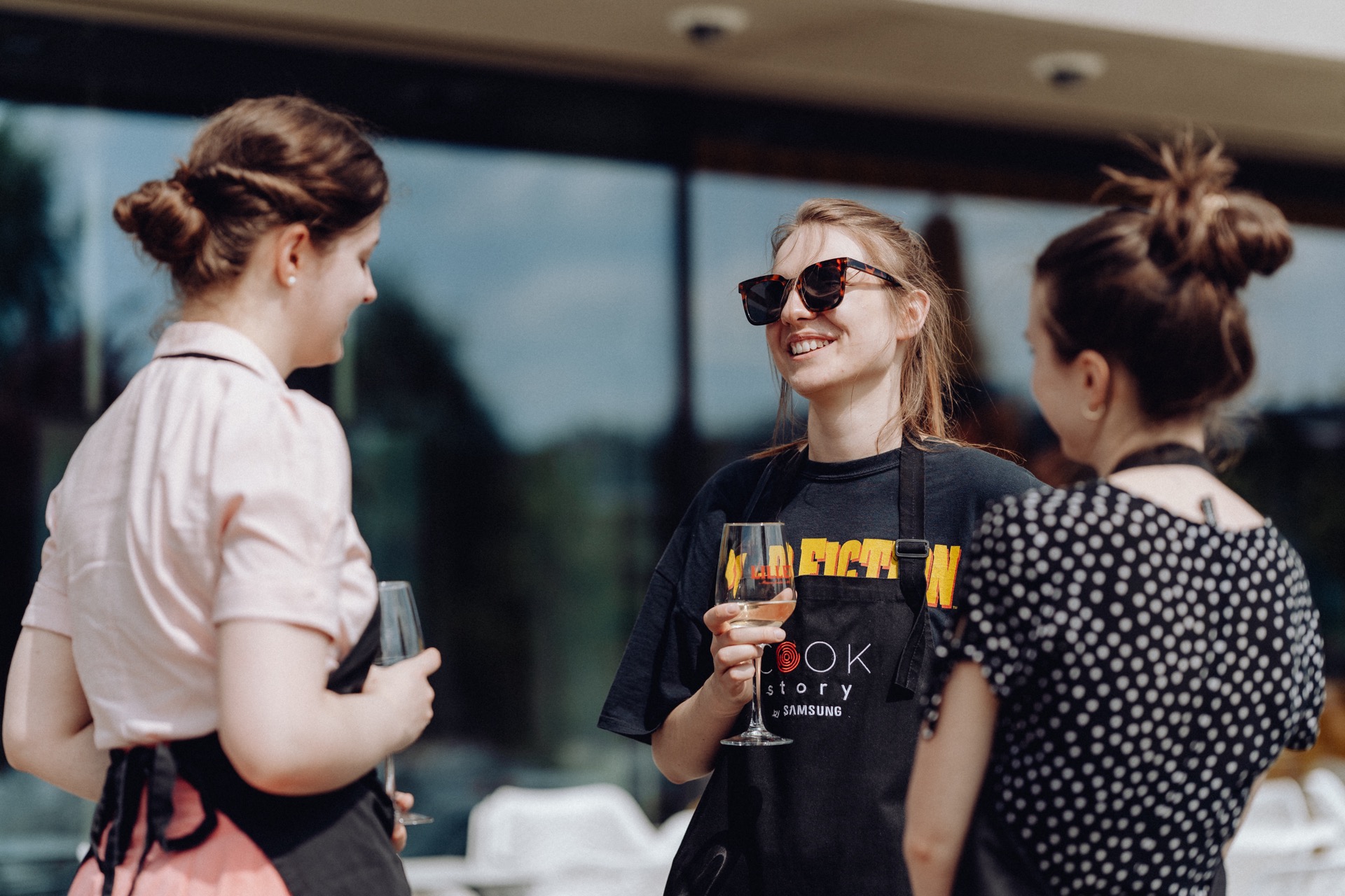 Three women standing outside, smiling and busy talking. They are wearing aprons and holding wine glasses. One woman is wearing sunglasses and a graphic T-shirt, while the others are dressed in bright tops and polka dots. The scene captures the essence of event photography against the backdrop of a building with large windows.   