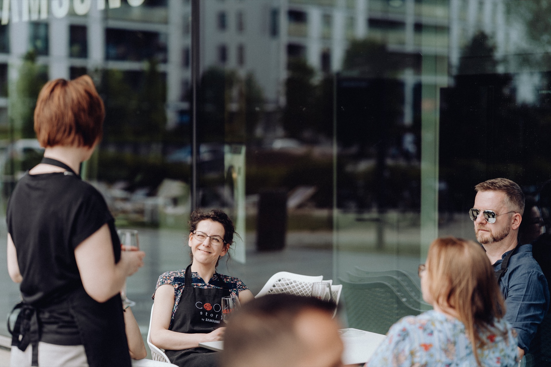 A group of people sitting at a table in an outdoor cafe, with one woman standing and serving drinks. The people sitting are talking and smiling, and the waiter has a glass of water in her hand. In the background are the reflective windows of the building - perfect for an event photo shoot.  