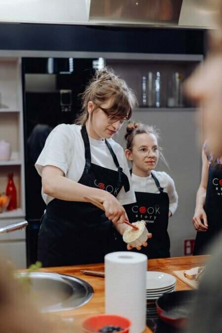 Dwie osoby ubrane w białe koszule i czarne fartuchy z logo „Cook Story” przygotowują jedzenie w kuchni. Osoba po lewej smaruje coś na kawałku chleba, a druga osoba patrzy. W tle widoczne są różne przedmioty kuchenne, co stanowi doskonałą fotorelację z wydarzeń.