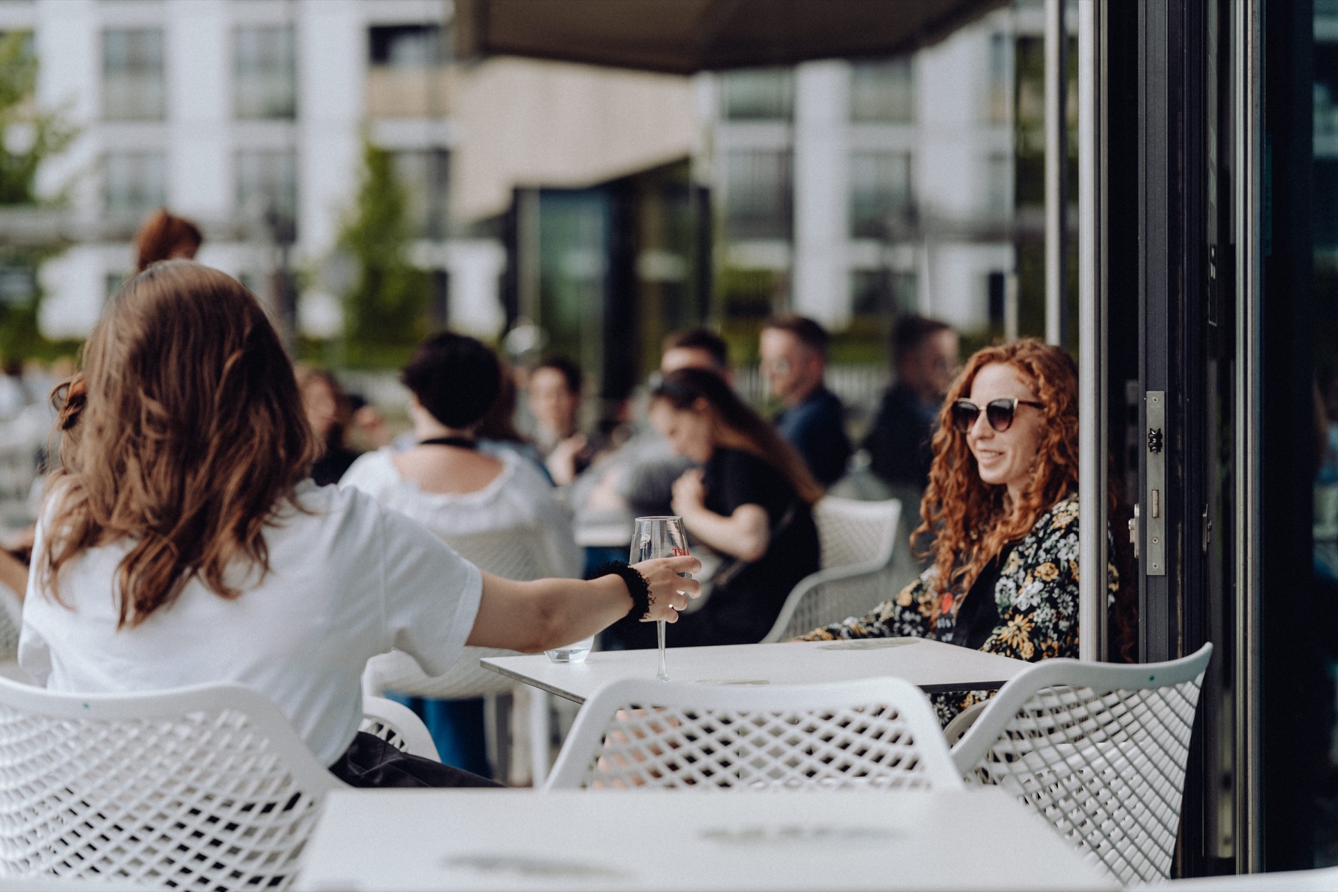 People are sitting outside in a cafe, socializing, chatting and drinking drinks. Attention focuses on a woman with curly red hair and sunglasses, sitting at a table to the right. Another woman in a white shirt, her back turned, raises her glass. The background is blurred, capturing the perfect moment of the photo event.   