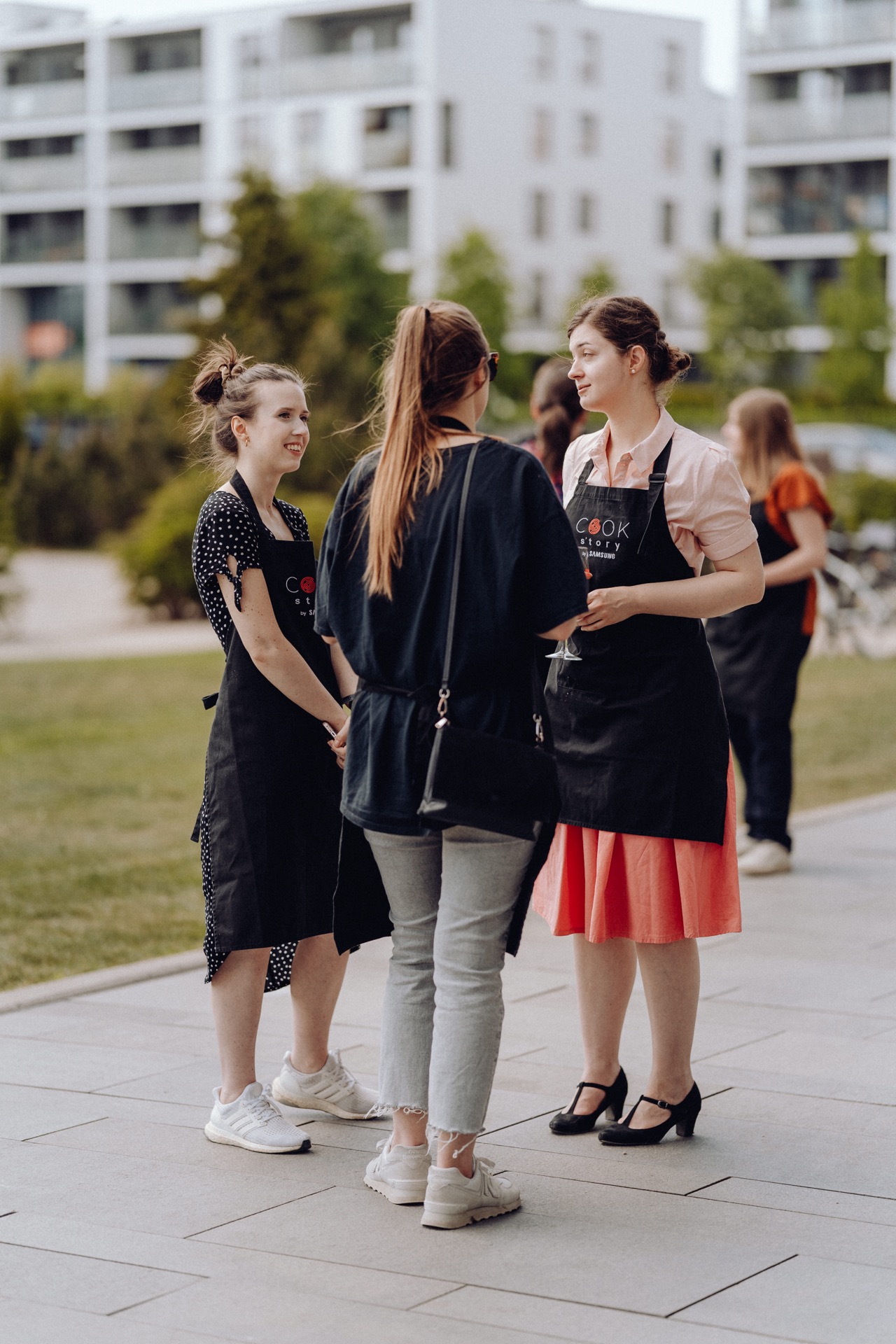 Three women stand outside and carry on a conversation. Two face the camera, dressed in black aprons and casual attire. The third, dressed in a coral dress and apron, is facing away. They look as if they are taking part in an outdoor event with people and buildings in the background - a perfect photo-op.   