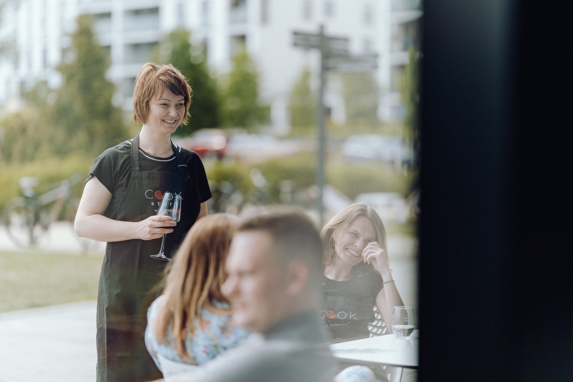 A smiling woman in a black apron holds a glass, standing next to an outdoor table. Two women sitting at the table are talking, one covers her mouth laughing. This is a charming event photography that shows the joy of the moment, with fuzzy people and greenery in the background.  