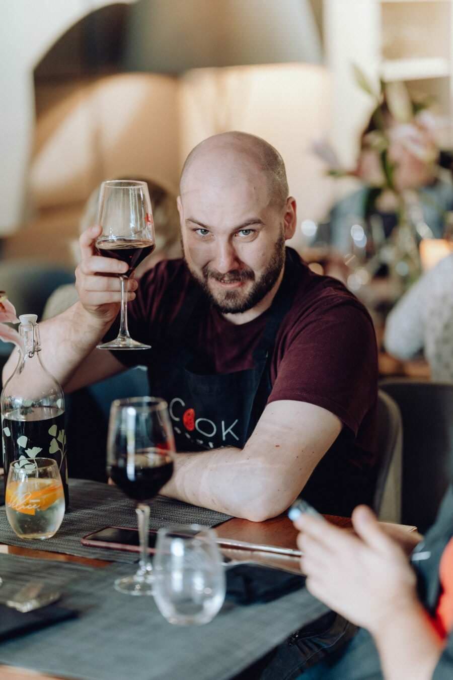 Mężczyzna w bordowej koszuli i czarnym fartuchu siedzi przy stole w restauracji, trzyma kieliszek czerwonego wina i uśmiecha się do kamery. Na stole znajdują się różne przedmioty, w tym szklana butelka z kwiatowym wzorem i smartfon na pierwszym planie, idealne do fotografii eventowej.