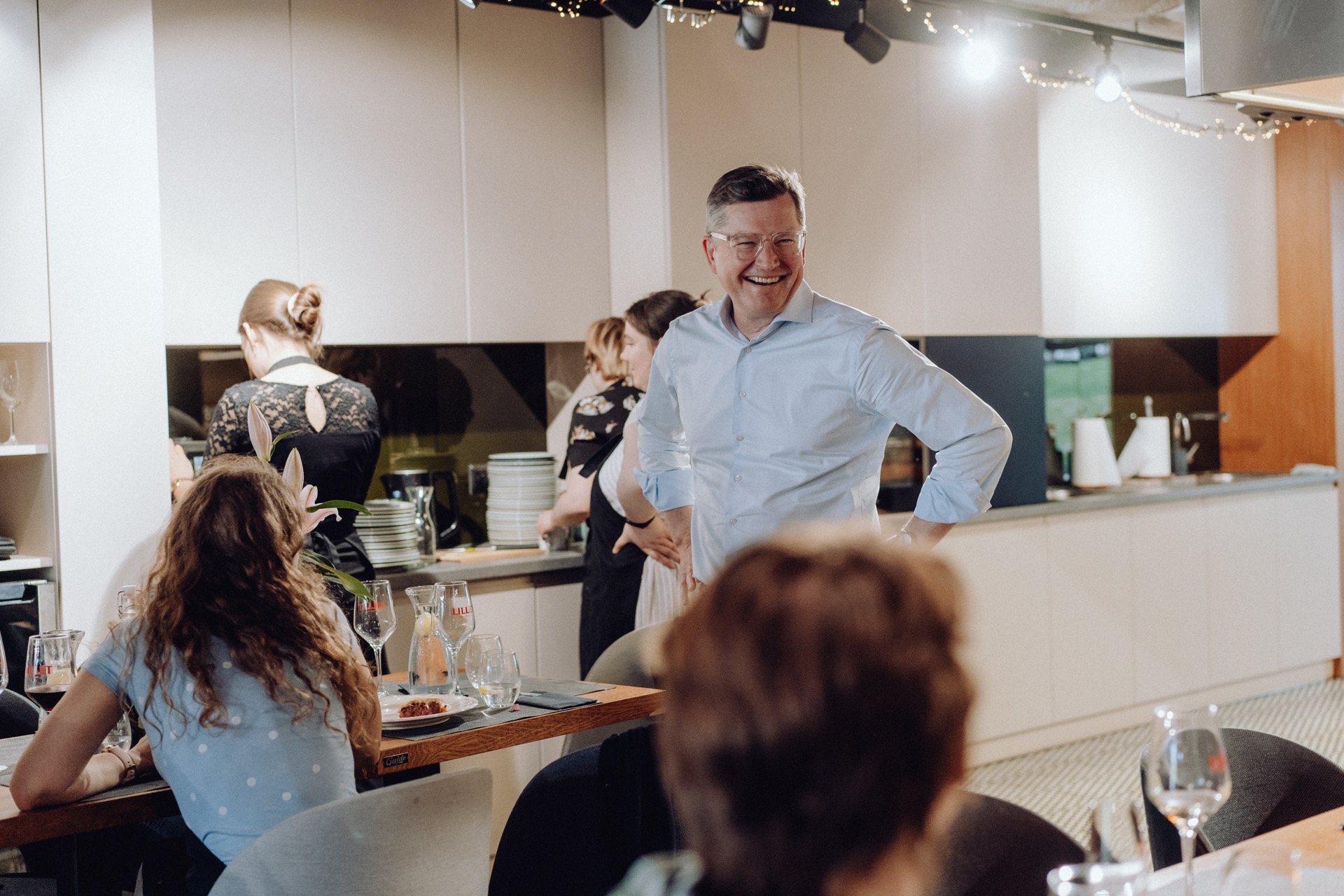 A man in a light blue shirt stands smiling with his hand on his hip in a modern kitchen. In the background, people are busy washing dishes and cleaning up. In the foreground, a child and an elderly person sit at a table set with plates, glasses and a bottle - it's like an inviting photo-reportage of events.  