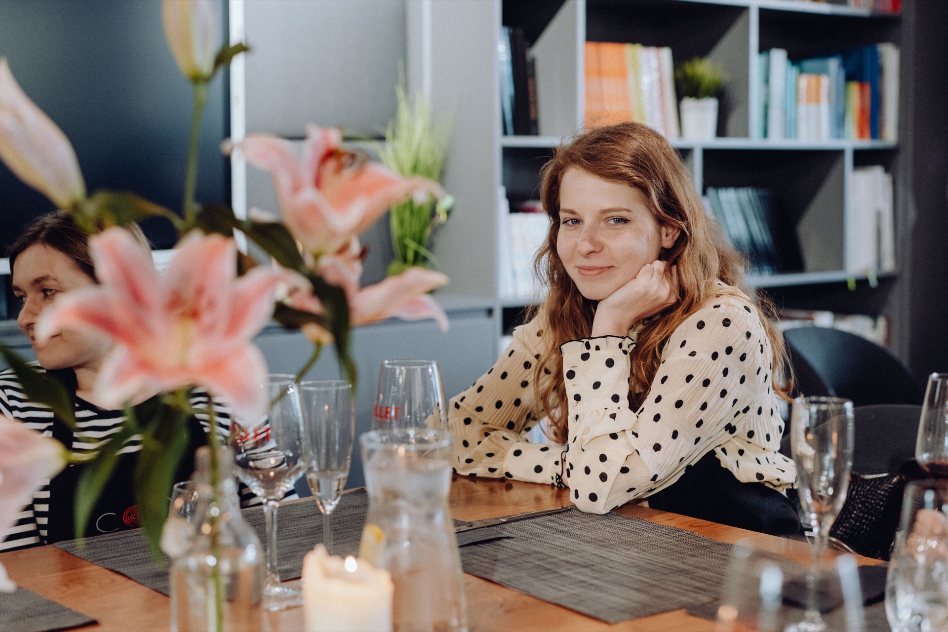 A woman with long red hair, wearing a polka dot blouse, sits at a table decorated with glass dishes, candles and pink flowers. Resting her chin on her hand and smiling, she radiates joy. Beside her, against a background of bookshelves, another person is partially visible - a perfect shot for event photography.  