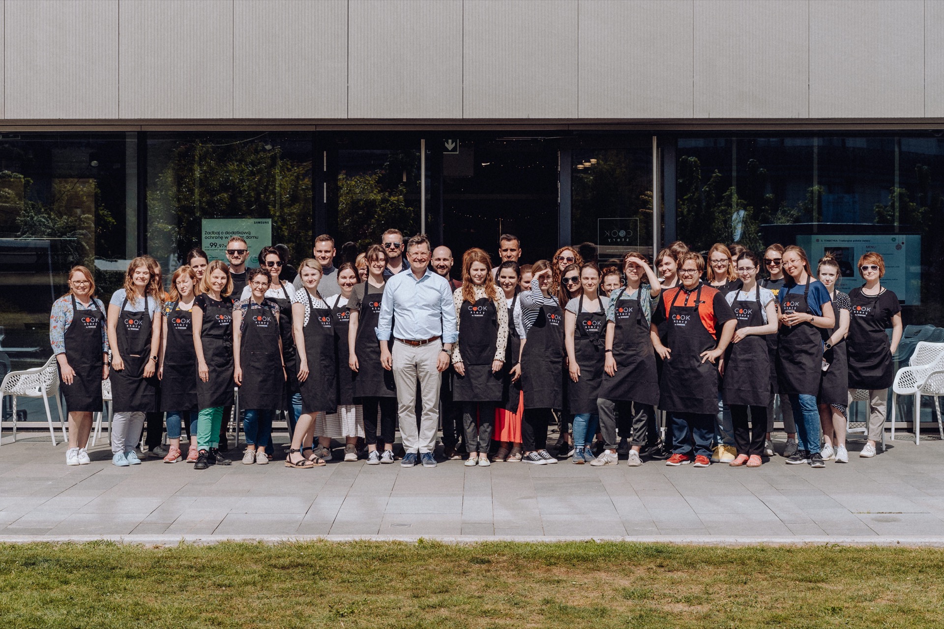 A large group of people standing in front of the building, all wearing black aprons with logos. They sit in several rows and smile at the camera. The area is paved and there is grass in front of the group - a perfect shot for event photography.  