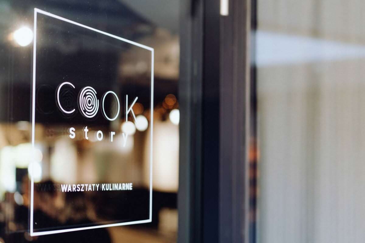 Zbliżenie na szklane drzwi z napisem „COOK Story” i „Warsztaty Kulinarne”. Przestrzeń wewnętrzna uchwycona przez fotografię eventową ma rozmyte tło z ciepłym oświetleniem, co sugeruje przytulne i przyjazne otoczenie.