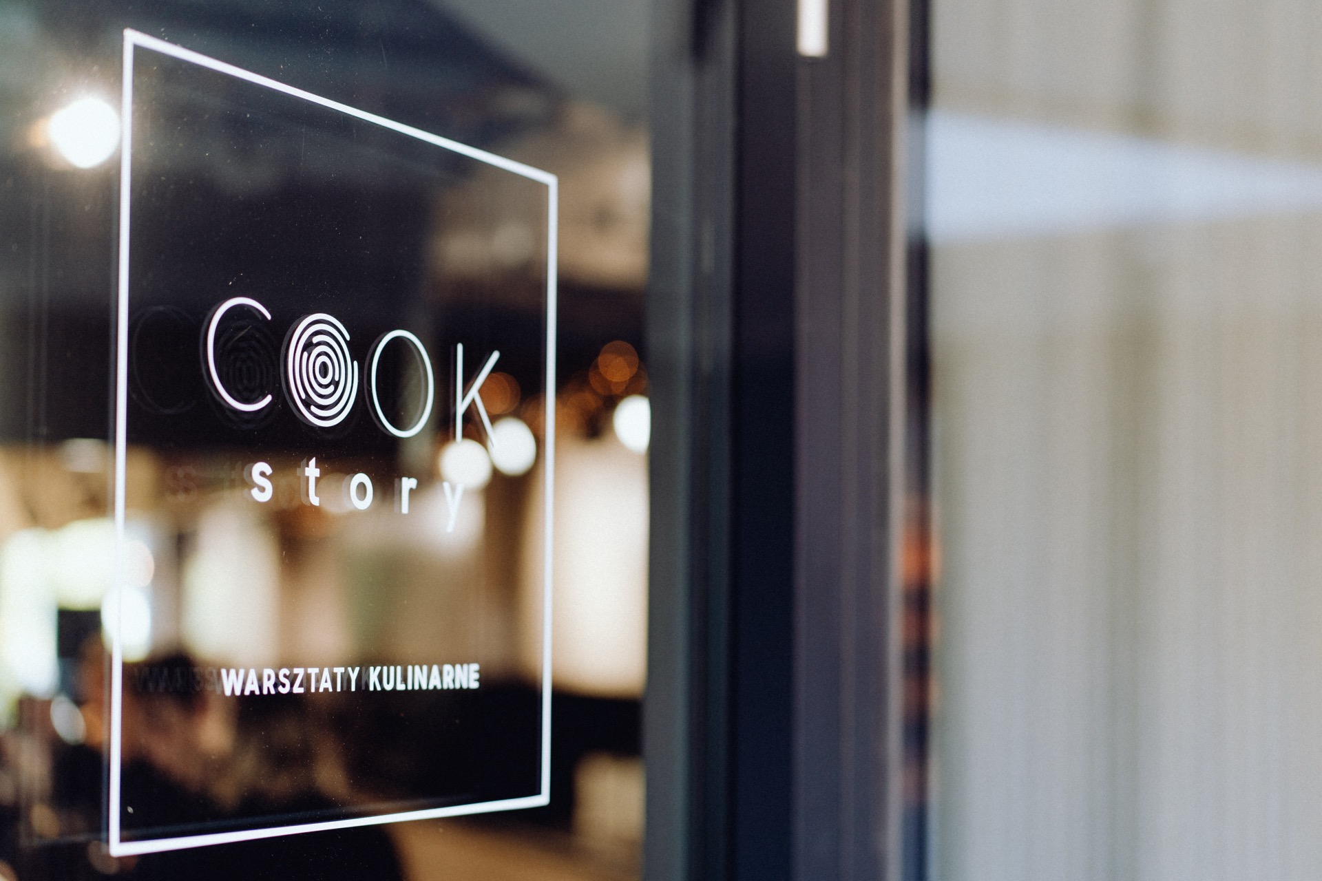 Close-up of a glass door with the words "COOK Story" and "Culinary Workshop." The interior space captured by the event photography has a blurred background with warm lighting, suggesting a cozy and friendly environment. 