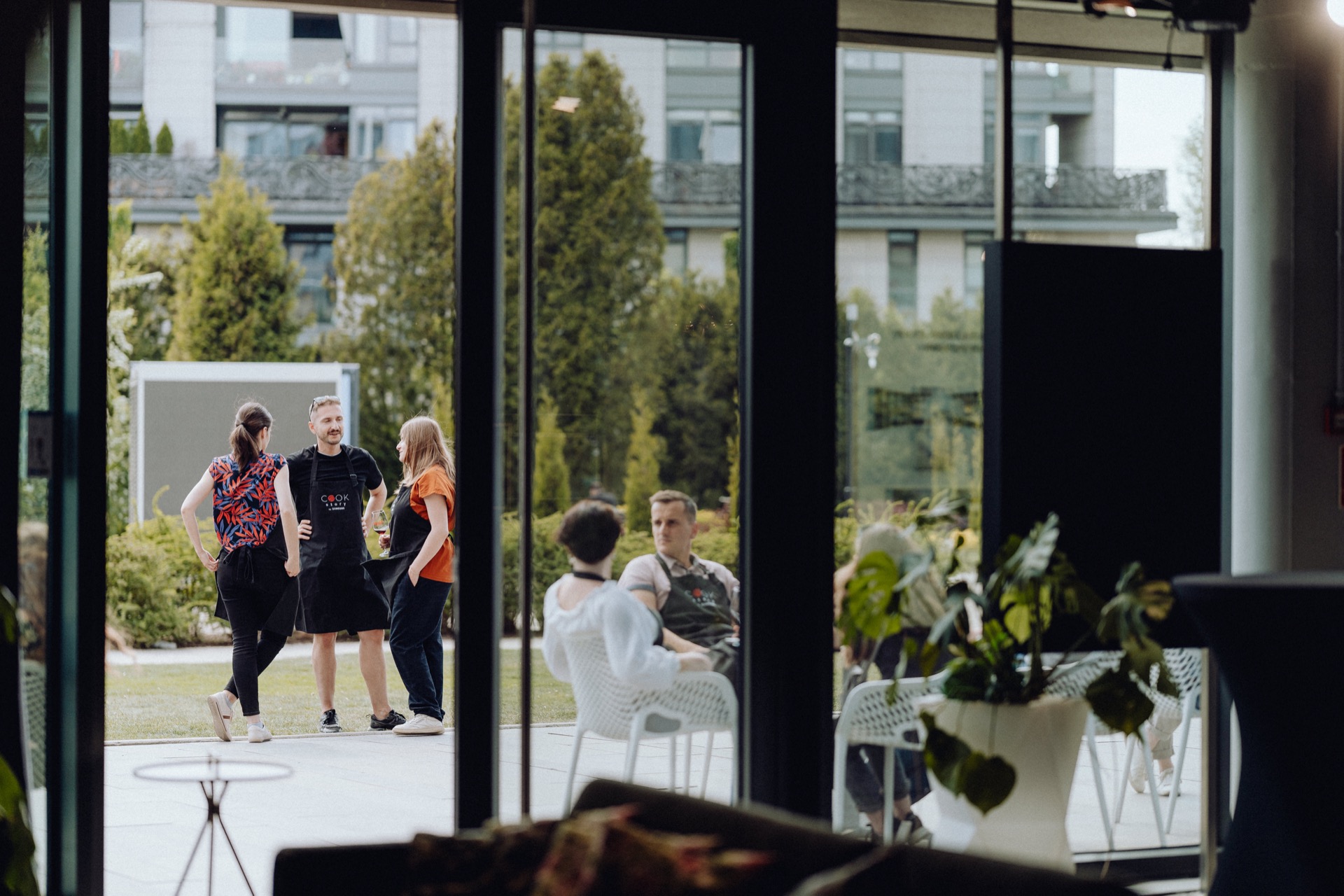 People meet outside, viewed through a glass door. Three people are standing and talking in the foreground, while two others are sitting on garden chairs in the background. The backdrop appears to be a modern building with greenery and plants, which the event photography captures beautifully.  