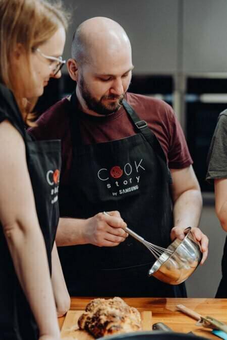 Mężczyzna ubrany w czarny fartuch „Cook Story” ubija składniki w metalowej misce. Dwie kobiety, również w fartuchach, stoją obok niego i przyglądają się. Ugotowane danie stoi przed nimi na drewnianym stole. Wydają się znajdować w nowoczesnej kuchni, pięknie uchwycone w ramach sesji fotograficznej eventowej.