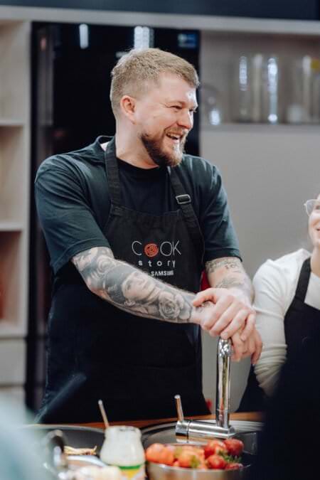 Osoba z tatuażami na ramionach, ubrana w czarny fartuch z napisem „Cook Story”, stoi w kuchni i uśmiecha się. Opierają się o blat, na którym leżą porozrzucane owoce i narzędzia kuchenne. Ten moment, doskonale uchwycony w ramach fotorelacji z wydarzeń, ukazuje obok nich częściowo widoczną inną osobę.