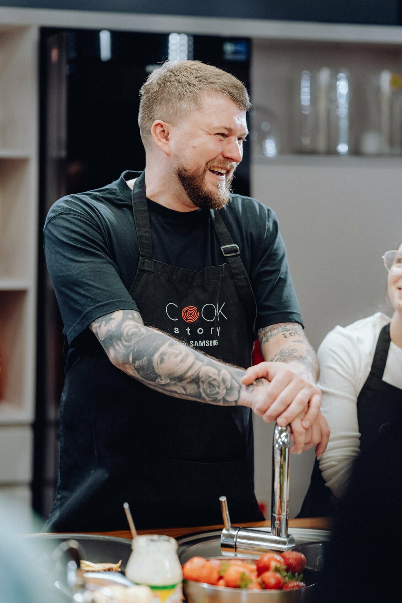 A person with tattoos on his arms, wearing a black apron with "Cook Story" written on it, stands in the kitchen and smiles. They are leaning against a countertop on which lie scattered fruits and kitchen tools. This moment, perfectly captured as part of a photo essay of the events, shows another person partially visible next to them.  