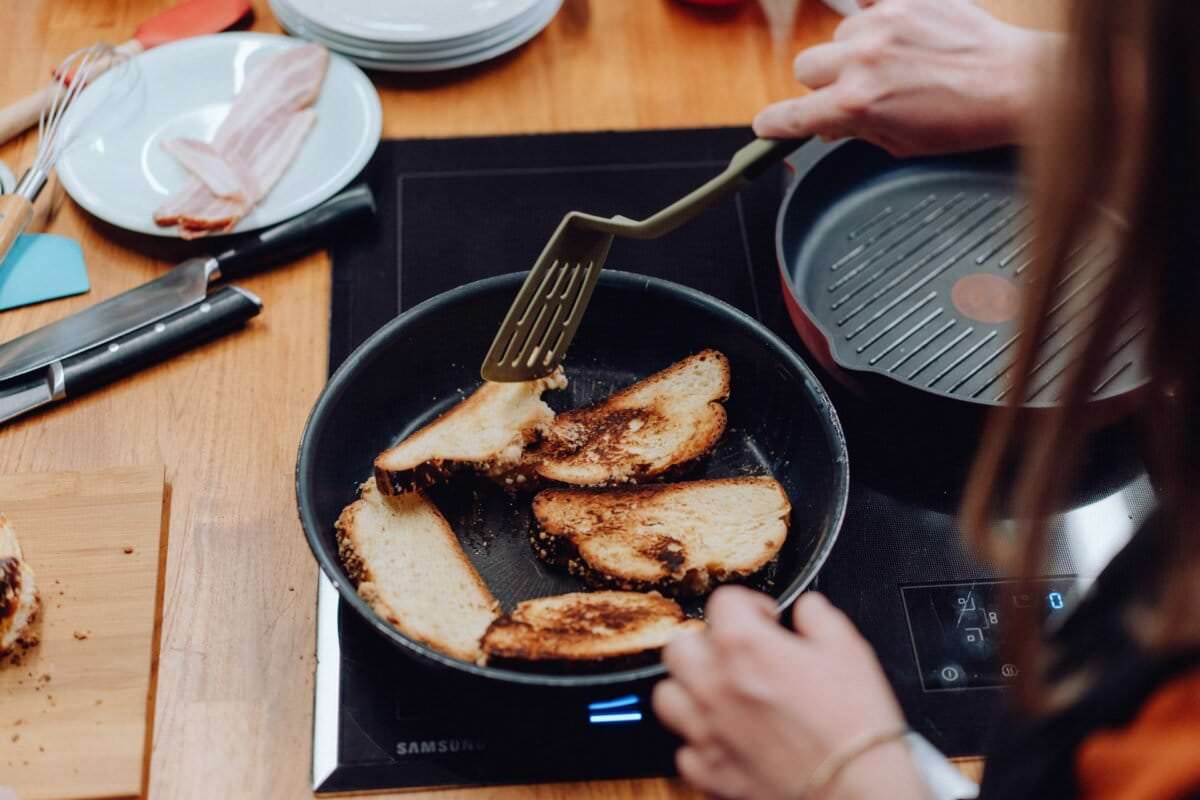 Widzi się osobę opiekającą kromki chleba na patelni nad płytą kuchenną. W pobliżu znajdują się starannie ułożone przybory kuchenne, deska do krojenia z większą ilością chleba, talerze i plastry boczku, oddając esencję komfortu domowej kuchni. Może to z łatwością stanowić część zachęcającej fotorelacji z wydarzeń w przytulnej kuchni.