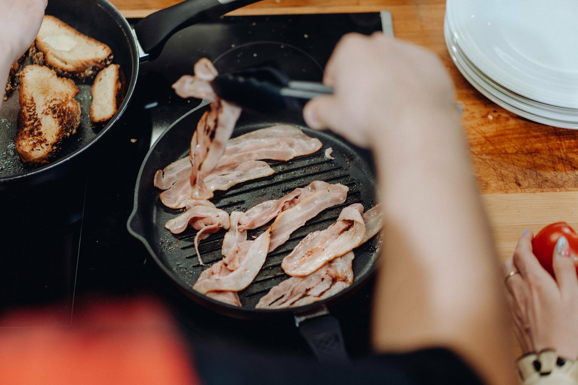 A person is frying bacon strips in a pan on the stove, capturing the moment like an event photographer warsaw. To the left, another pan with toasted bread can be seen. A person holds tongs, while a tomato and a stack of plates adorn the right side of a wooden countertop, creating a picturesque photo of the event.  