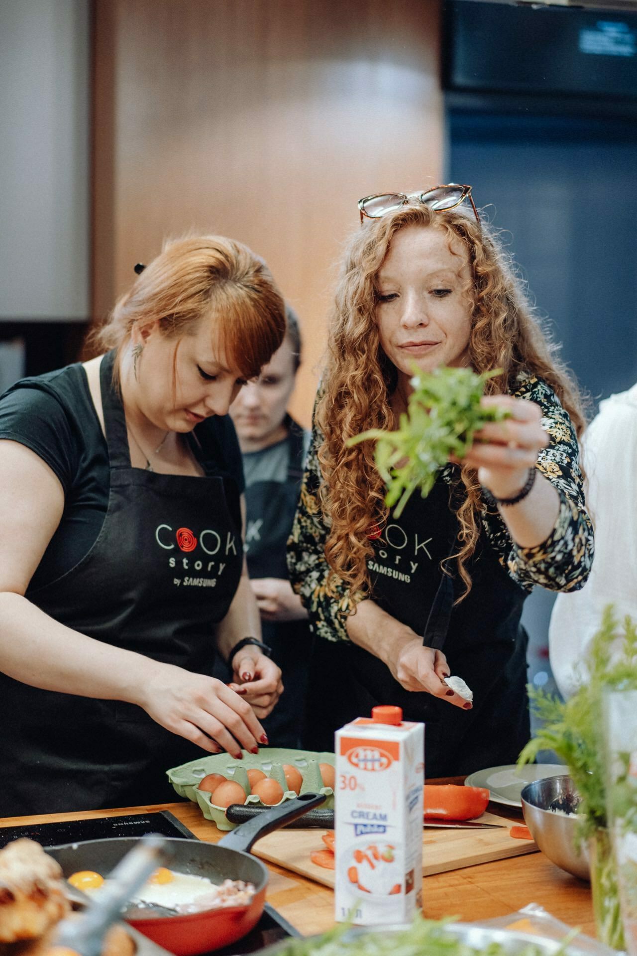 Two women dressed in black aprons with "Cook Story" logos are preparing food in the kitchen. One woman puts vegetables into a dish, while the other with curly hair holds fresh herbs. A carton of cream and various ingredients lie on a wooden countertop - a perfect scene for event photography.  