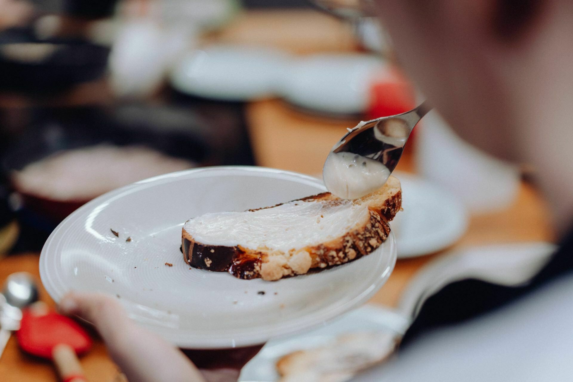 A person spreads a creamy paste on a slice of bread with a butter knife. The bread rests on a white plate, which the person holds along with the knife. Additional kitchen tools and ingredients are blurred in the background, capturing an intimate moment worthy of an event photo essay.  