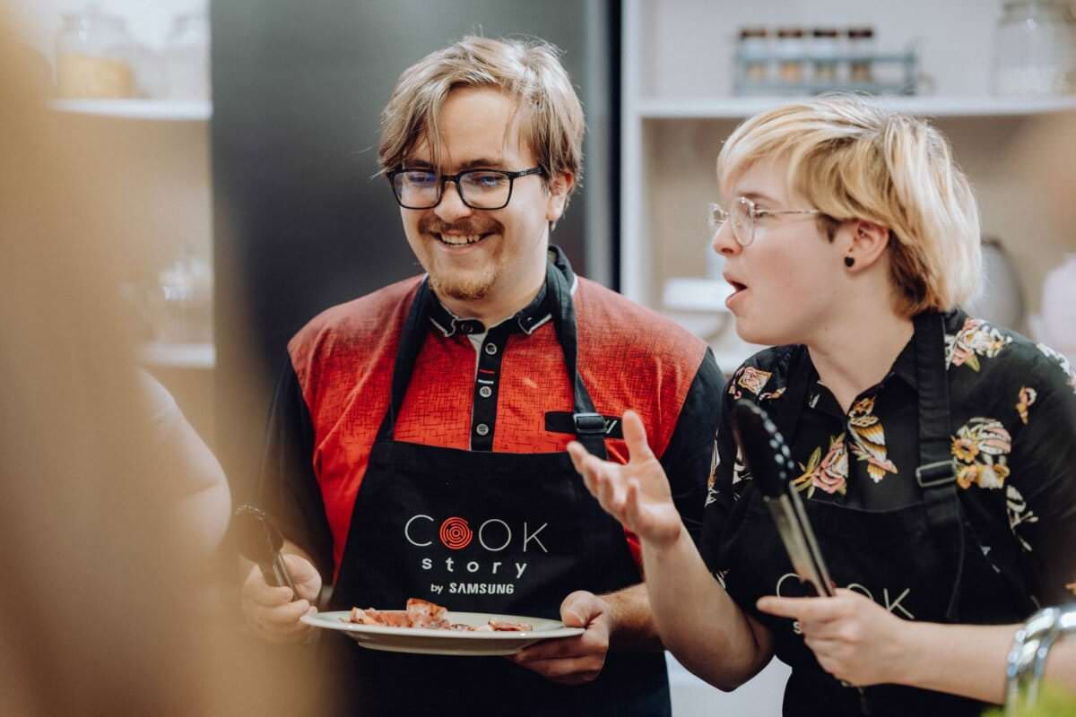 Dwie osoby ubrane w fartuchy kuchenne biorą udział w zajęciach kulinarnych. Osoba po lewej stronie uśmiecha się trzymając talerz z jedzeniem, a osoba po prawej gestykuluje z ożywieniem podczas mówienia. W tle widoczne przybory kuchenne i półki ze słoikami, idealne do fotografii eventowej.