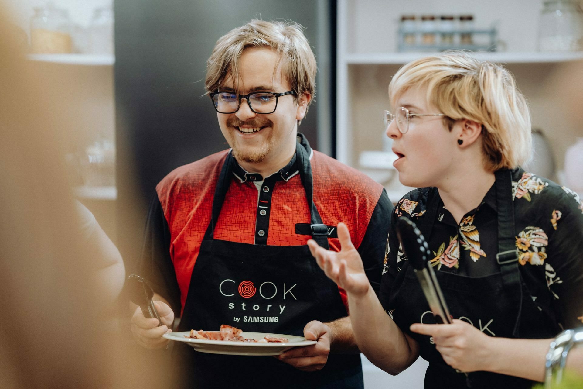 Two people dressed in kitchen aprons take part in a cooking activity. The person on the left is smiling while holding a plate of food, and the person on the right is gesturing animatedly while speaking. Kitchen utensils and shelves of jars are visible in the background, perfect for event photography.  