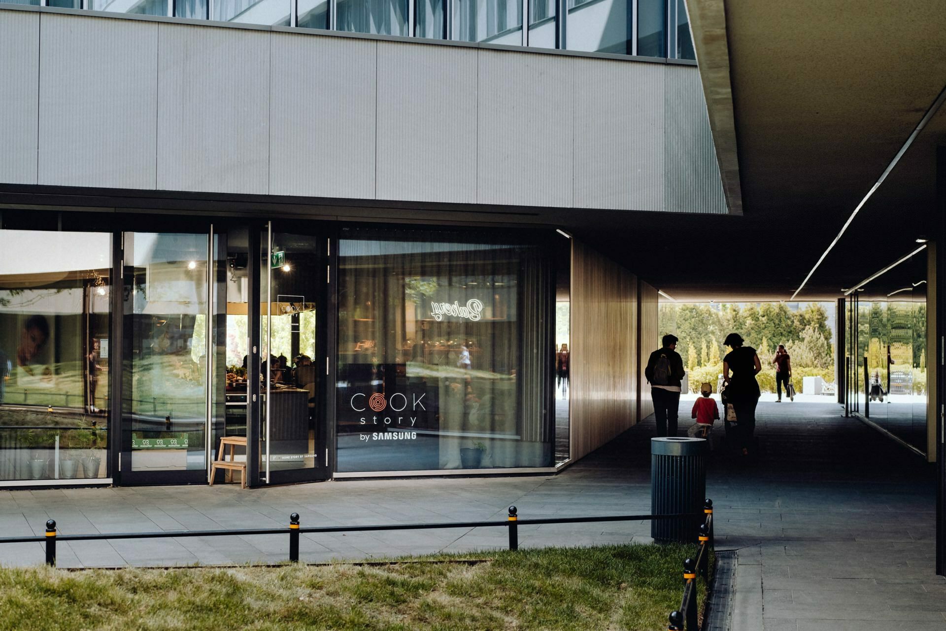 The modern building features large glass storefronts that read "COOK Story by Samsung." People walk along a shaded corridor next to the building, leading to a sunny open space with greenery visible in the background - an ideal location for event photography. 