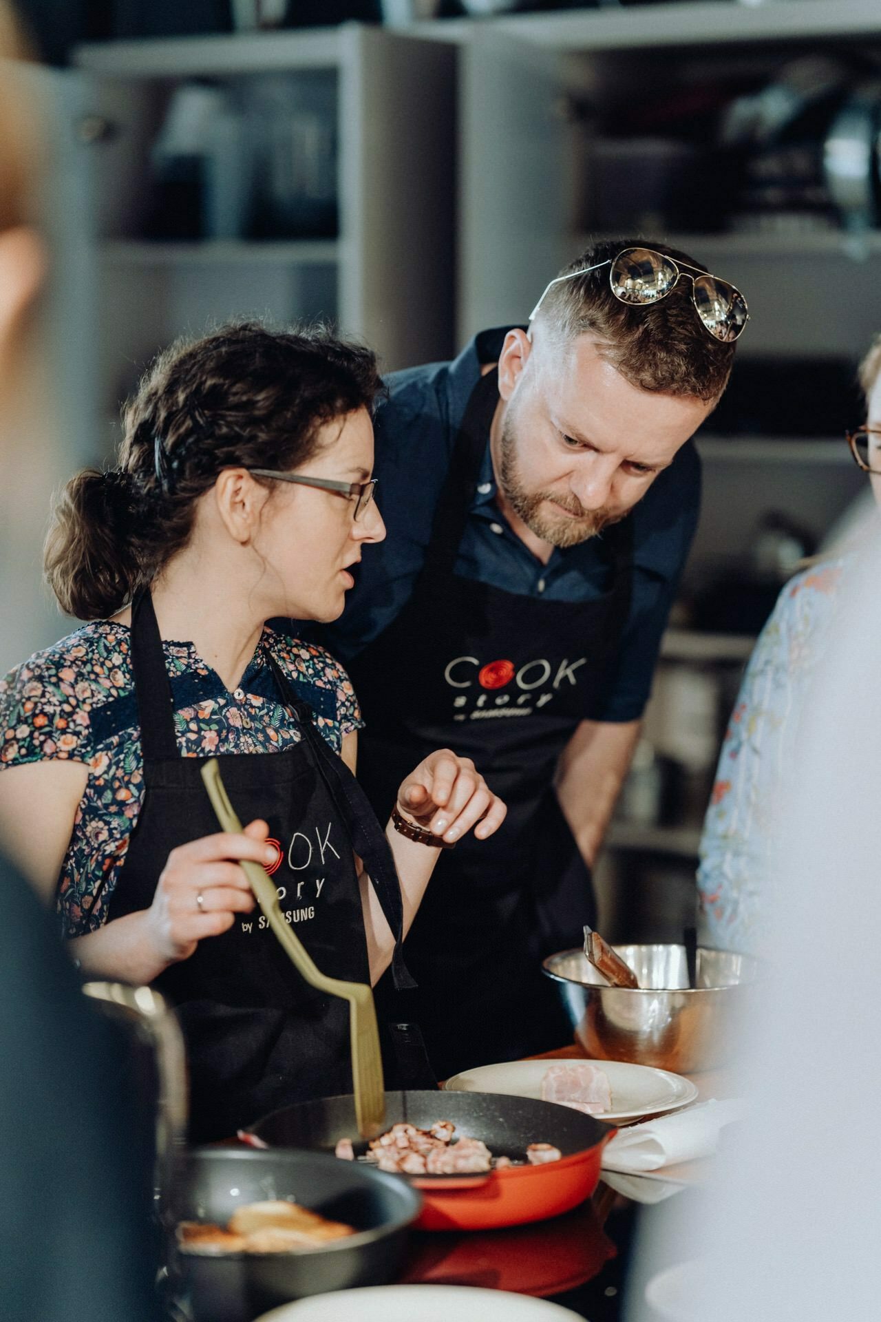 Two people, a man and a woman, are engaged in cooking in the kitchen. Both are wearing black aprons with the word "COOK" written on them. The woman stirs the food in the pan, while the man, wearing sunglasses on his head, watches and listens intently to her - a perfect moment for any event photographer warsaw.  