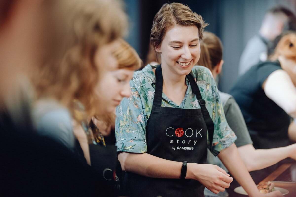 Kobieta ubrana w kwiecistą koszulę i fartuch z napisem „Cook Story by Samsung” uśmiecha się, uczestnicząc w lekcjach gotowania. Otaczają ją inni uczestnicy, którzy również skupiają się na zajęciach kulinarnych, tworząc żywą i wciągającą atmosferę, idealną do fotografii eventowej.