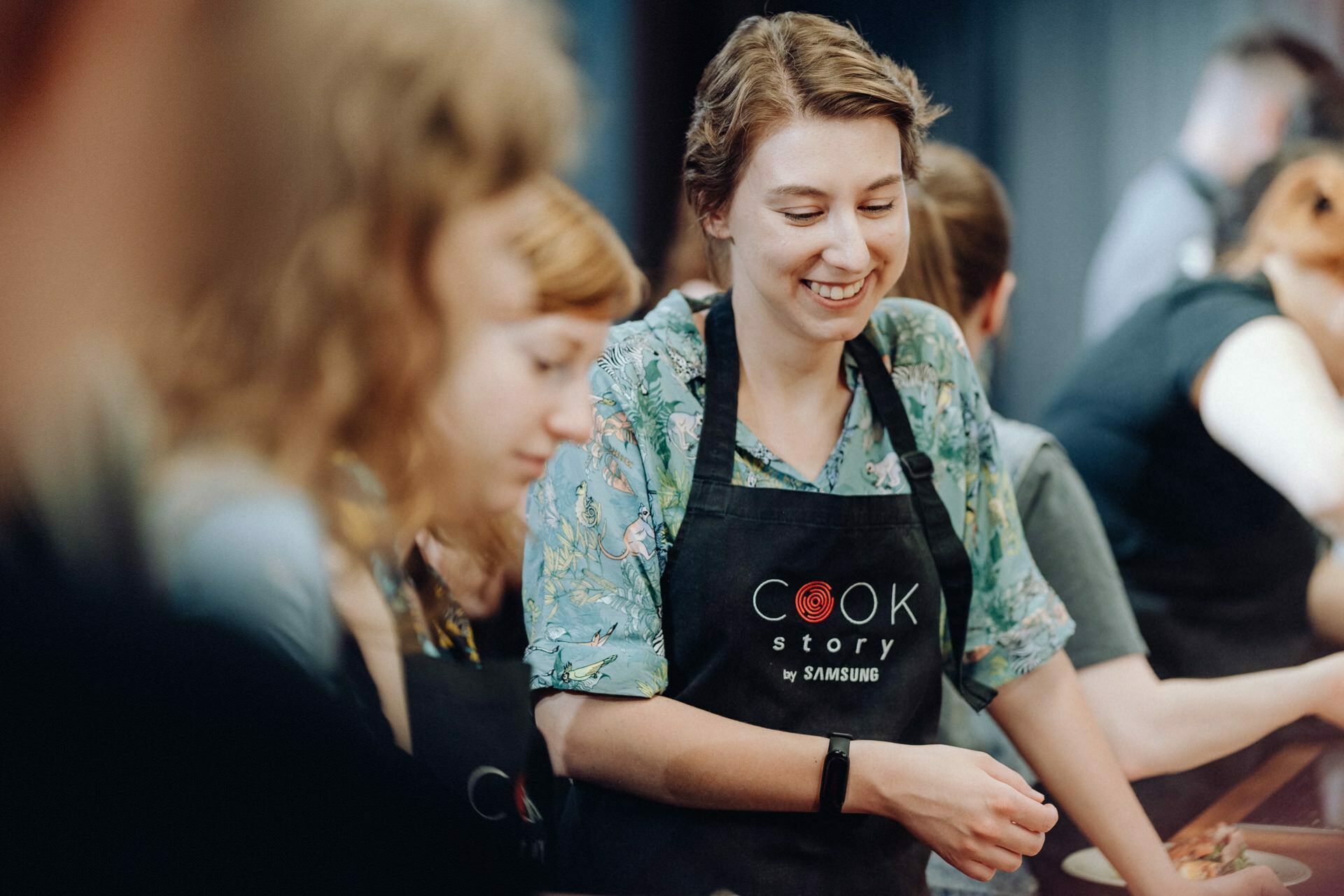 A woman dressed in a floral shirt and apron with the words "Cook Story by Samsung" smiles as she participates in a cooking class. She is surrounded by other participants who are also focused on the cooking class, creating a lively and engaging atmosphere, perfect for event photography. 