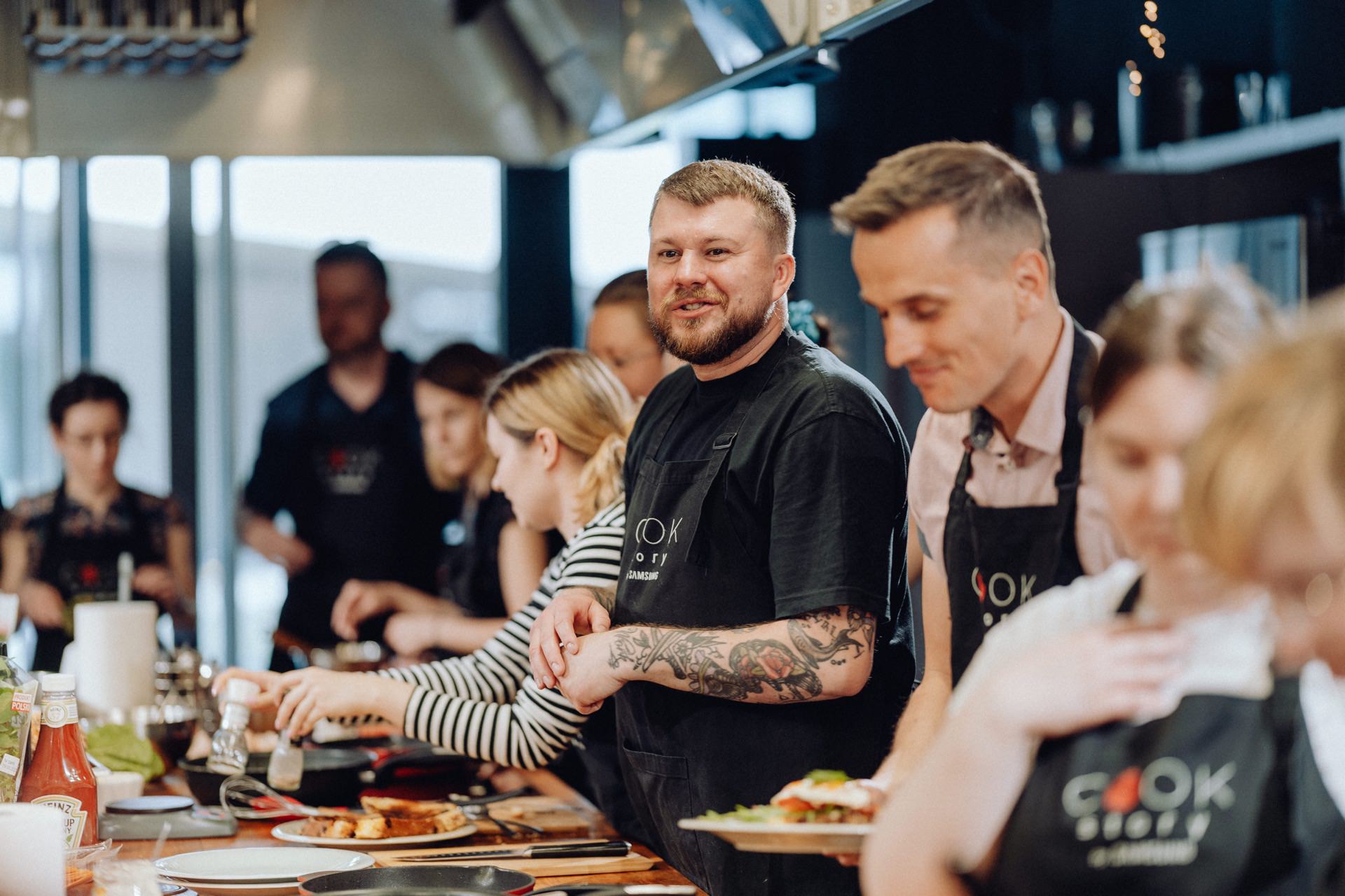 A group of people dressed in aprons participate in a cooking class in a modern kitchen. A man with tattoos and a black apron smiles in the middle, while others are busy preparing food around a long table with various ingredients for cooking. Perfect for an event photo shoot or event photoshoot!  