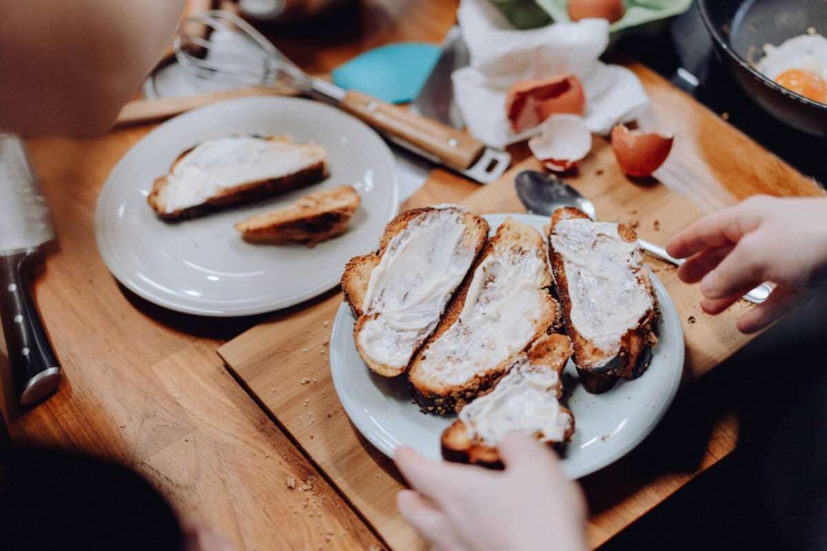 Widać dwie dłonie przygotowujące tosty na talerzu, każdą posmarowaną kremową pastą. Stół zawalony przyborami kuchennymi, potłuczonymi skorupkami jajek, nożem i trzepaczką, co sugeruje aktywne przygotowywanie posiłków. To tak, jakby uchwycenie chwili w fotorelacji z wydarzeń dodało dodatkowej warstwy autentyczności tej ruchliwej scenie.