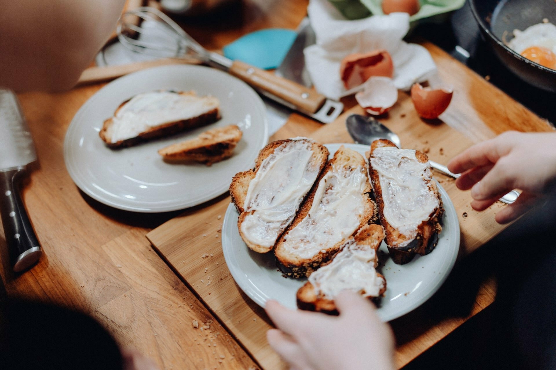 Two hands can be seen preparing toast on a plate, each spread with a creamy paste. The table is swamped with kitchen utensils, broken eggshells, a knife and a whisk, suggesting active food preparation. It's as if capturing the moment in a photo recap of events adds an extra layer of authenticity to this busy scene.  