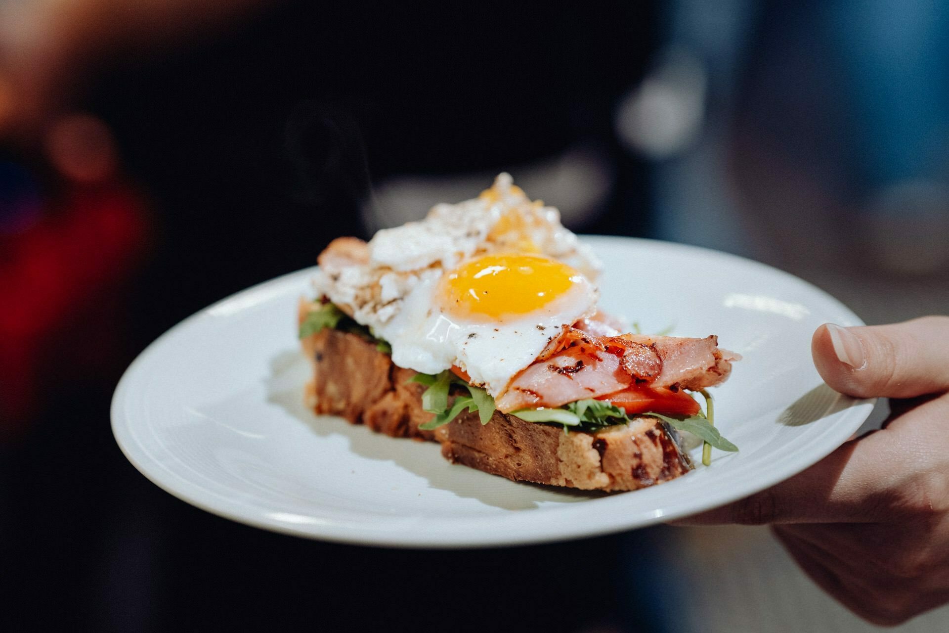 A person holds a white plate on which lies an open-faced sandwich, beautifully decorated with arugula, tomato slices, crispy bacon and an egg sunny side up with a liquid yolk. The background is blurred, capturing the essence of event photography. 