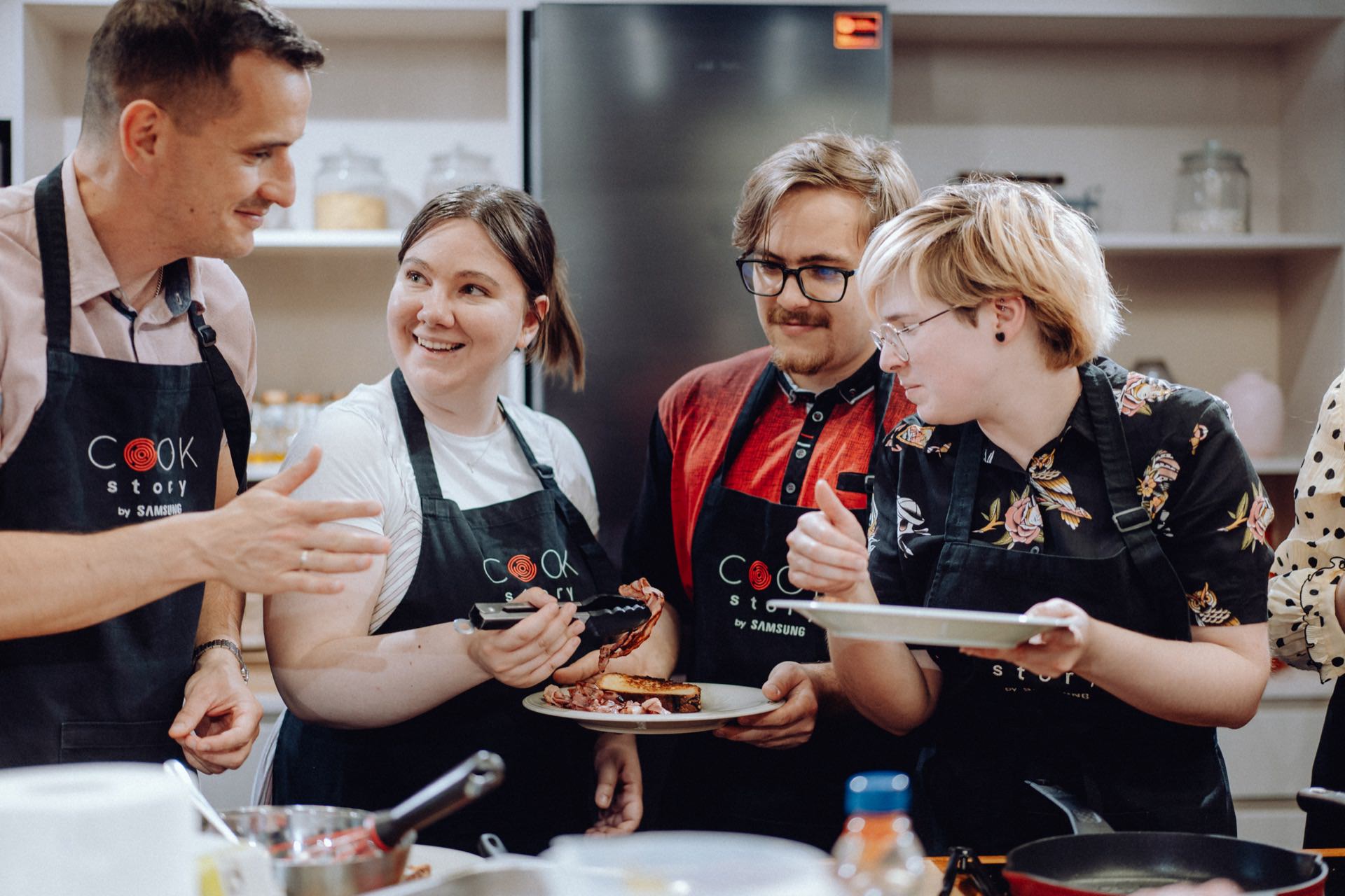 A group of four people in aprons laugh and engage while preparing food in the kitchen. Two hold plates of food, while the other focuses on adding the finishing touches. A refrigerator and kitchen shelves are visible in the background, perfectly captured for the event photo essay.  