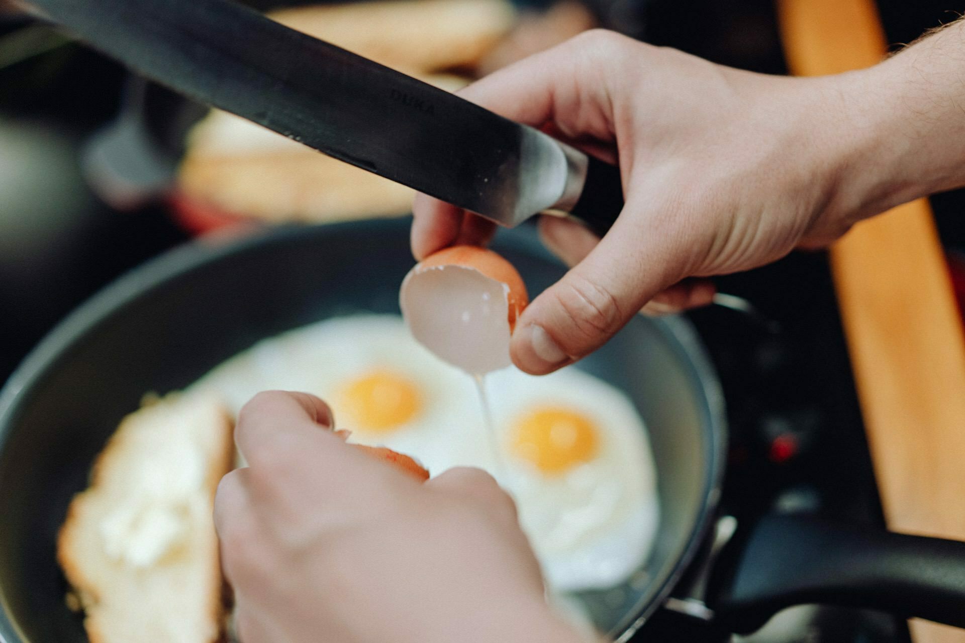 A close-up shows hands breaking an egg in a frying pan, while two fried eggs are already sizzling. The photo-like scene takes place in the kitchen, where a knife carefully assists in the action. 