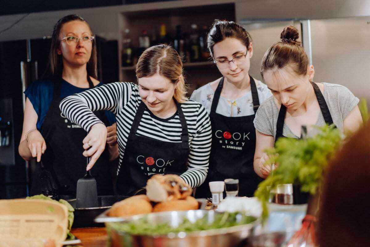 Grupa czterech kobiet biorących udział w lekcjach gotowania, wszystkie ubrane w czarne fartuchy z napisem „Cook Story”. Koncentrują się na przygotowywaniu i gotowaniu jedzenia. Składniki i przybory kuchenne porozrzucane są na blacie przed nimi, doskonale uchwycone jako część angażującego wydarzenia fotograficznego.