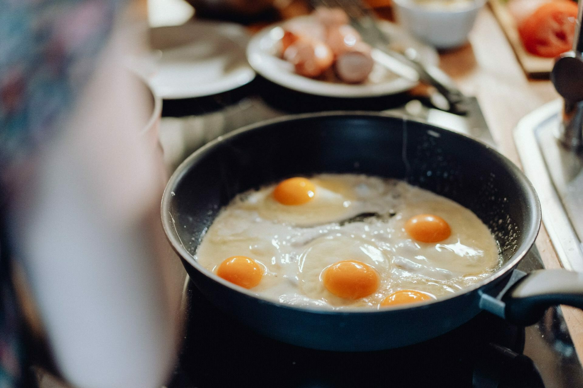 A close-up of a pan on the stove where several eggs are frying, some with yolks intact, others spread out. This shot could easily become part of an event photograph that captures the essence of a cozy morning. In the background are plates with sliced bread and various kitchen utensils.  