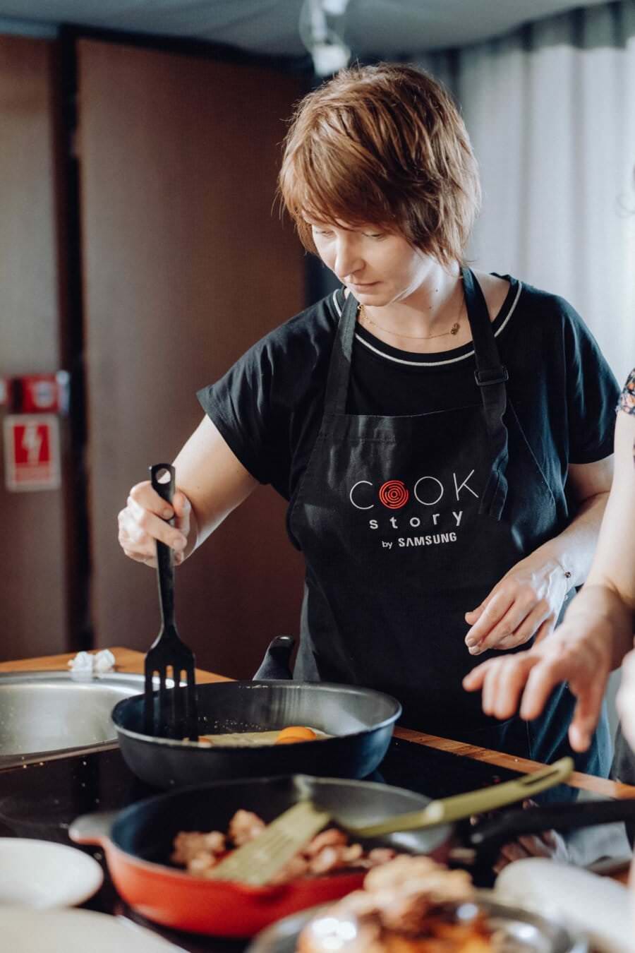 Kobieta ubrana w czarny fartuch z napisem „Cook Story by Samsung” gotuje w kuchni, uchwycona na fotografii eventowej. Używa szpatułki do mieszania jedzenia na patelni, podczas gdy po prawej stronie widoczna jest dłoń innej osoby trzymającej naczynie.