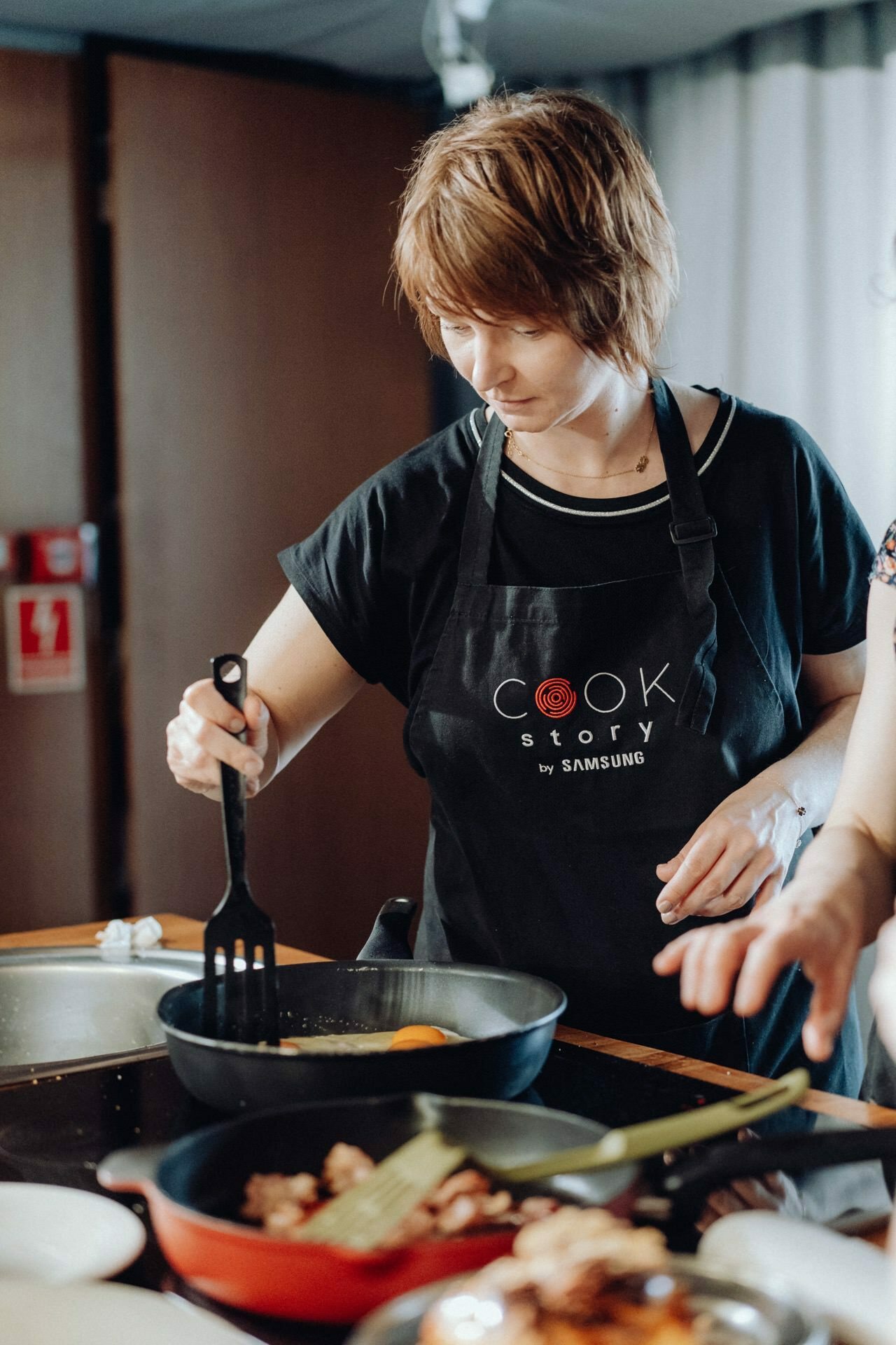 A woman wearing a black apron with the words "Cook Story by Samsung" is cooking in the kitchen, captured in an event photo. She is using a spatula to stir food in a pan, while to the right another person's hand can be seen holding a dish. 