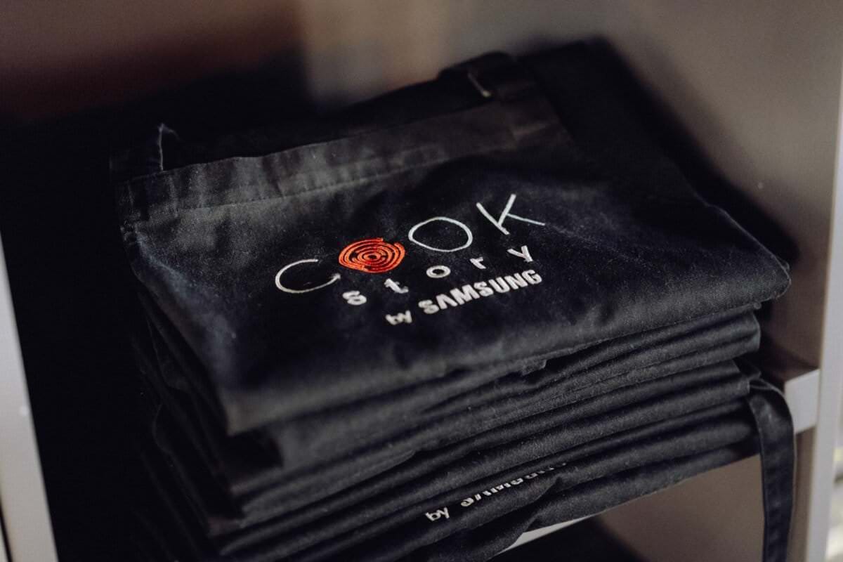 Na półce stoi starannie ułożony stos czarnych fartuchów z wyhaftowanym logo „Cook Story by Samsung”. Logo, wykonane przez eksperta fotografa eventowego Warszawa, zawiera słowo „Cook” z pomarańczowym wirem zintegrowanym z literą „O” oraz znajdujący się pod nim napis „Story by Samsung”.